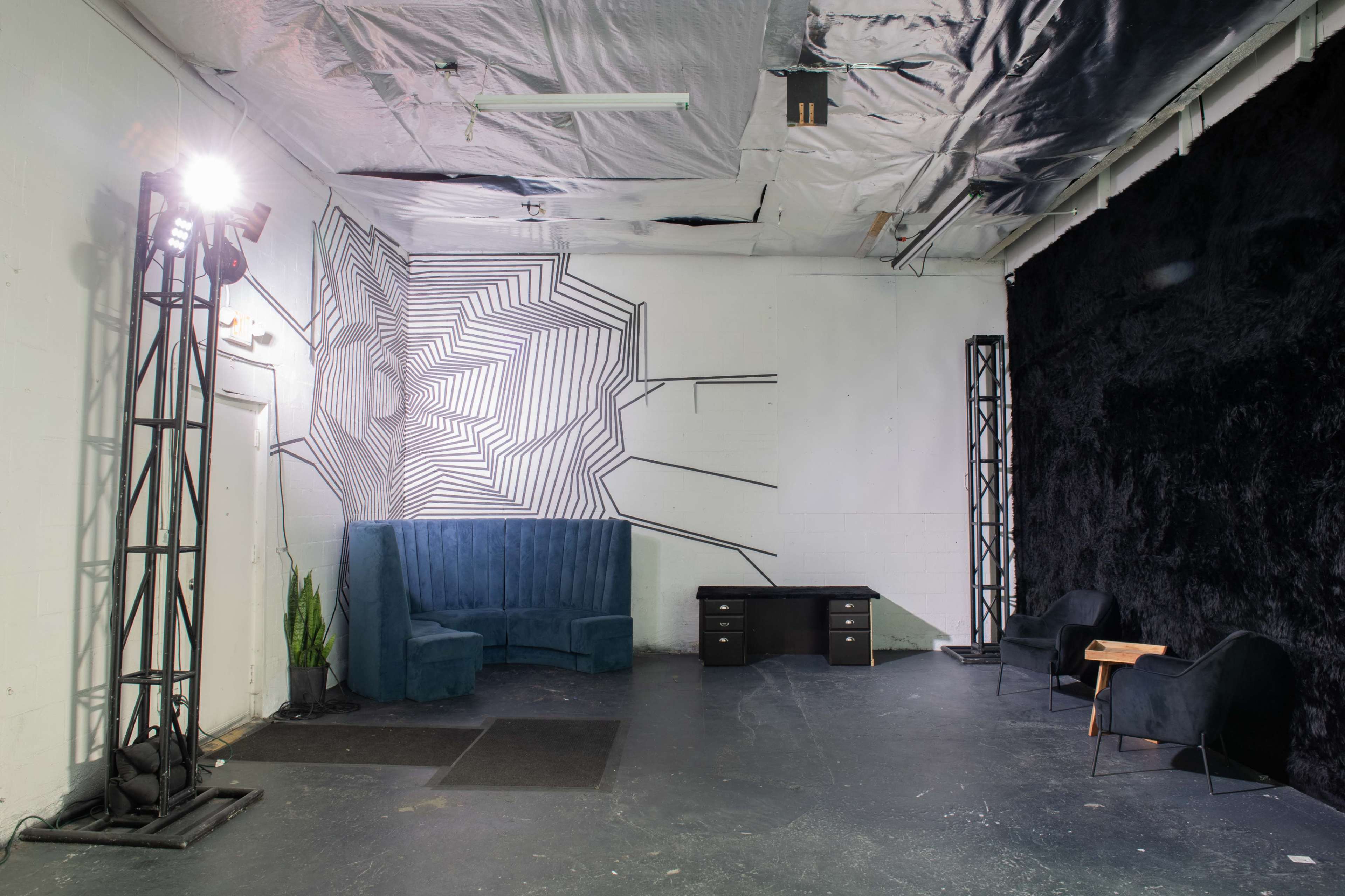 An empty studio space featuring a geometric wall design, a blue upholstered seating area, a black wall, and various furniture elements including desks and chairs.