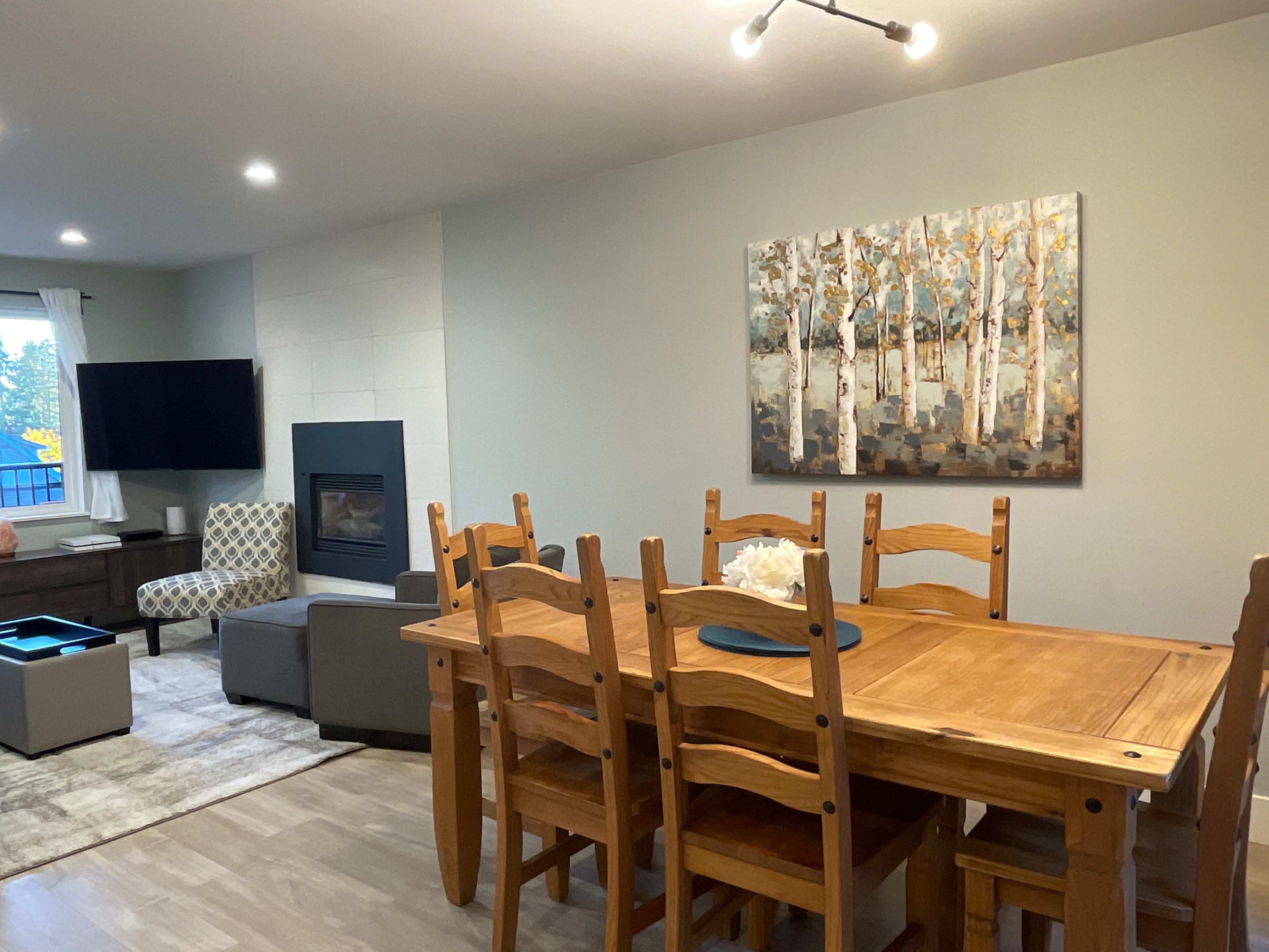 A wooden dining table with chairs is set in a room featuring a modern fireplace and a large painting of birch trees on the wall.