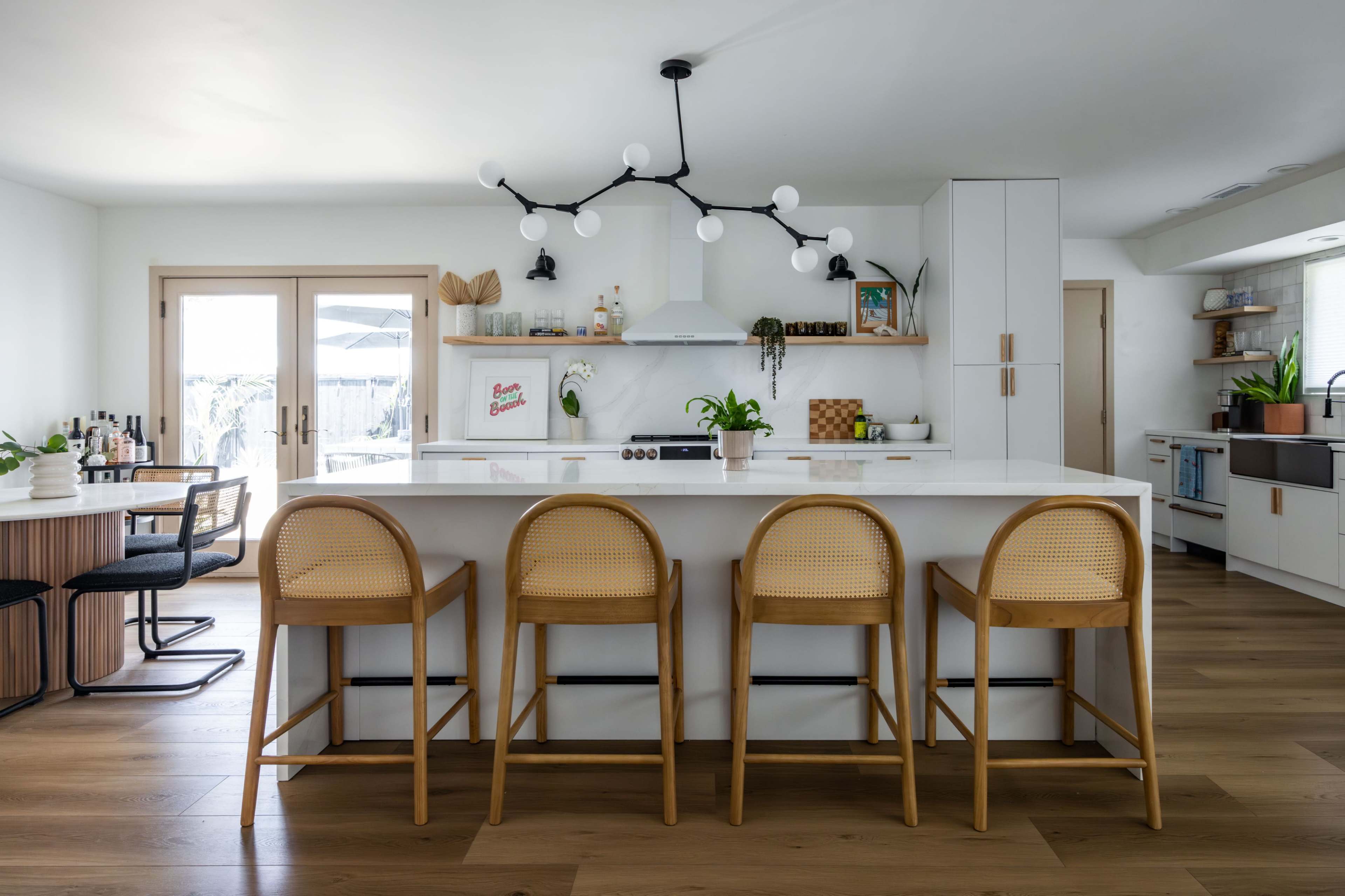 A modern kitchen features a large island with four rattan bar stools, white cabinetry, and plants, alongside wooden accents and a light fixture with multiple bulbs.