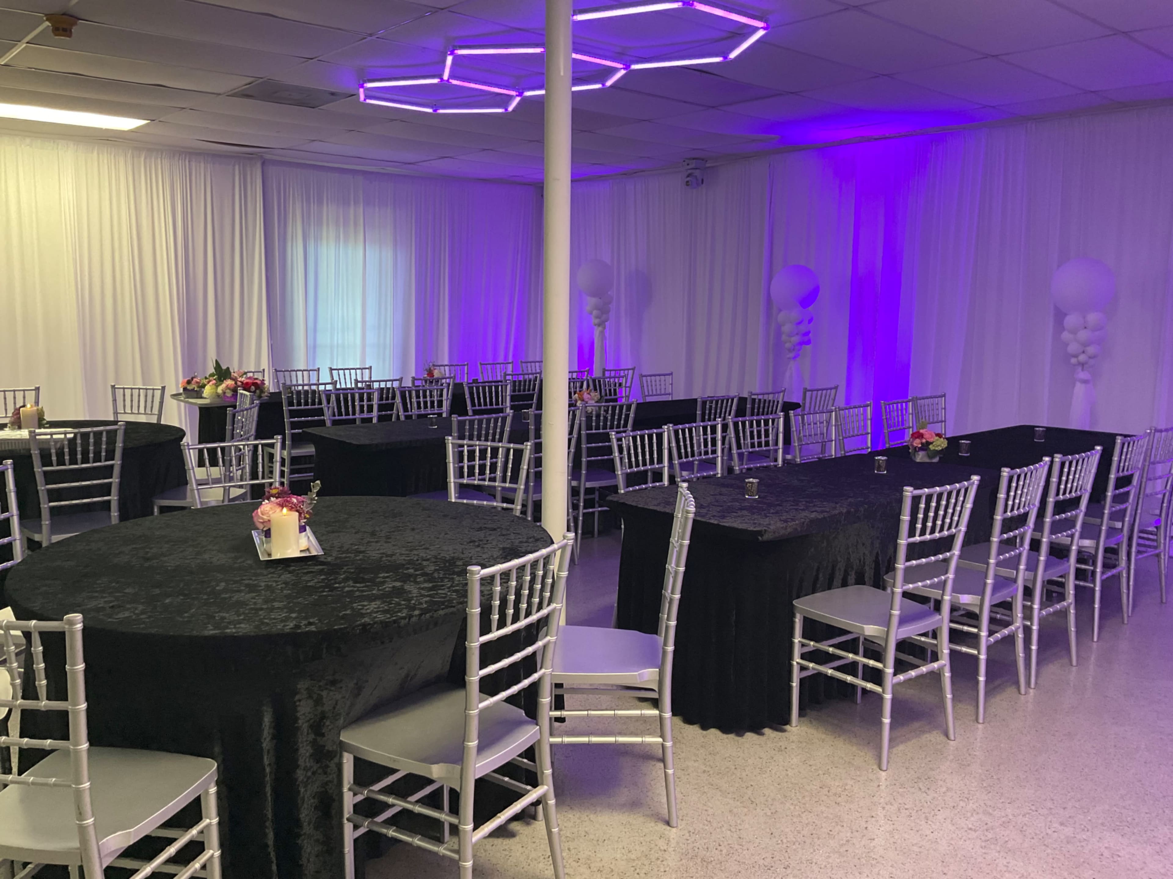 The image shows a decorated event space with round and rectangular tables covered in black tablecloths, silver chiavari chairs, and ambient purple lighting.