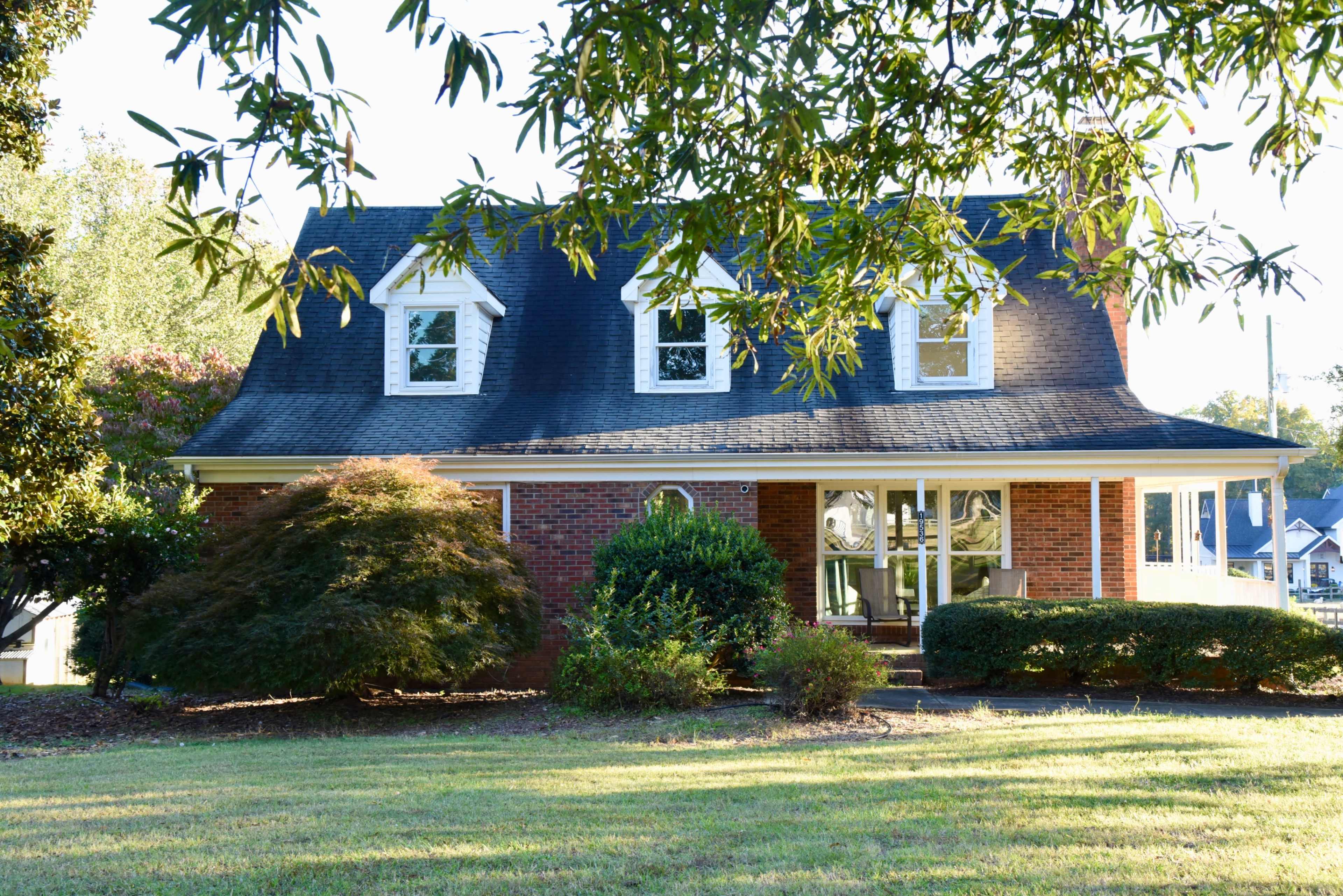 A red-brick house with a steep roof and white trim is surrounded by leafy shrubs and trees.