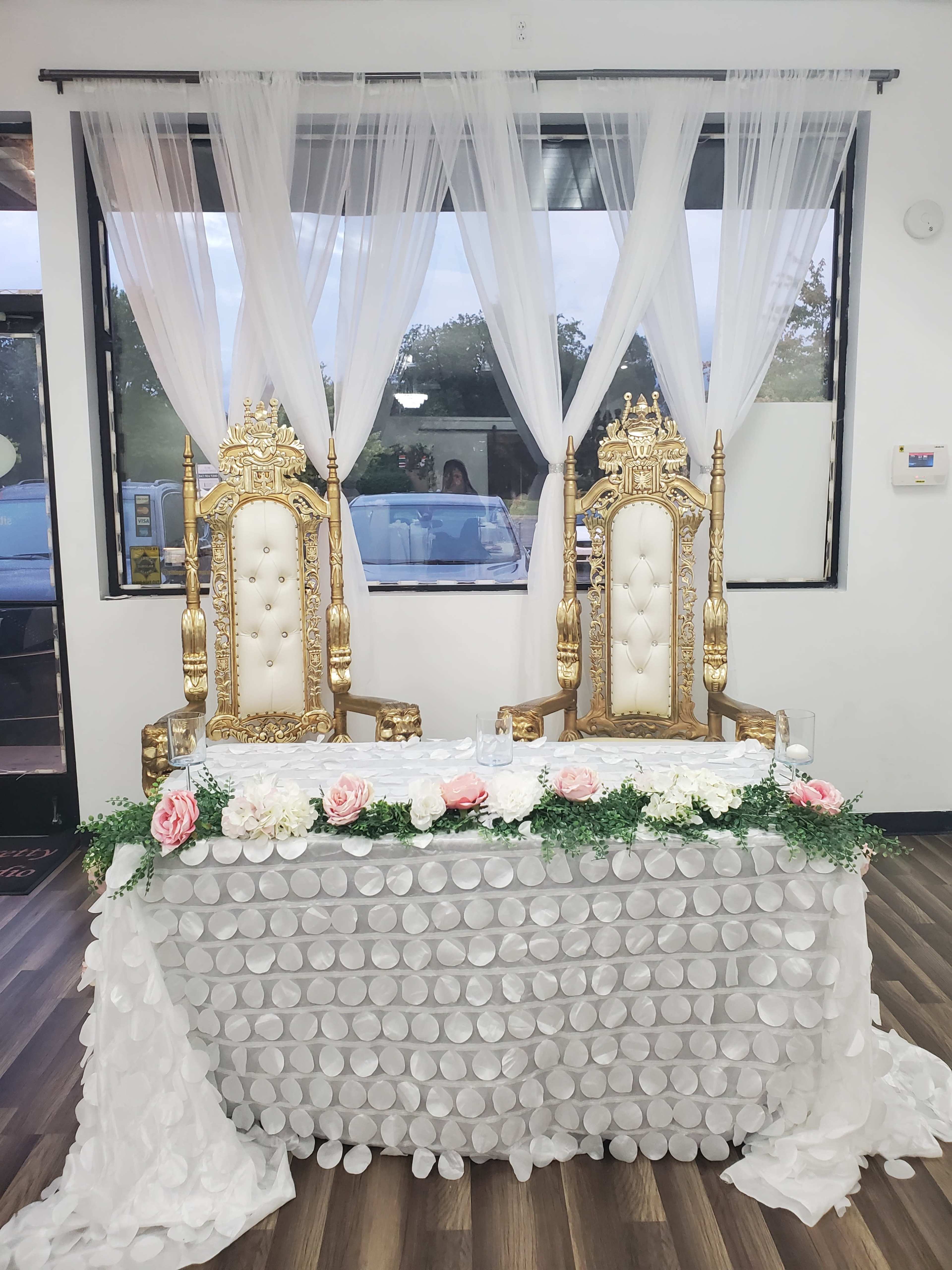 A decorative setup featuring two golden thrones placed on a table adorned with white fabric and floral arrangements, set against a backdrop of sheer curtains and a window.