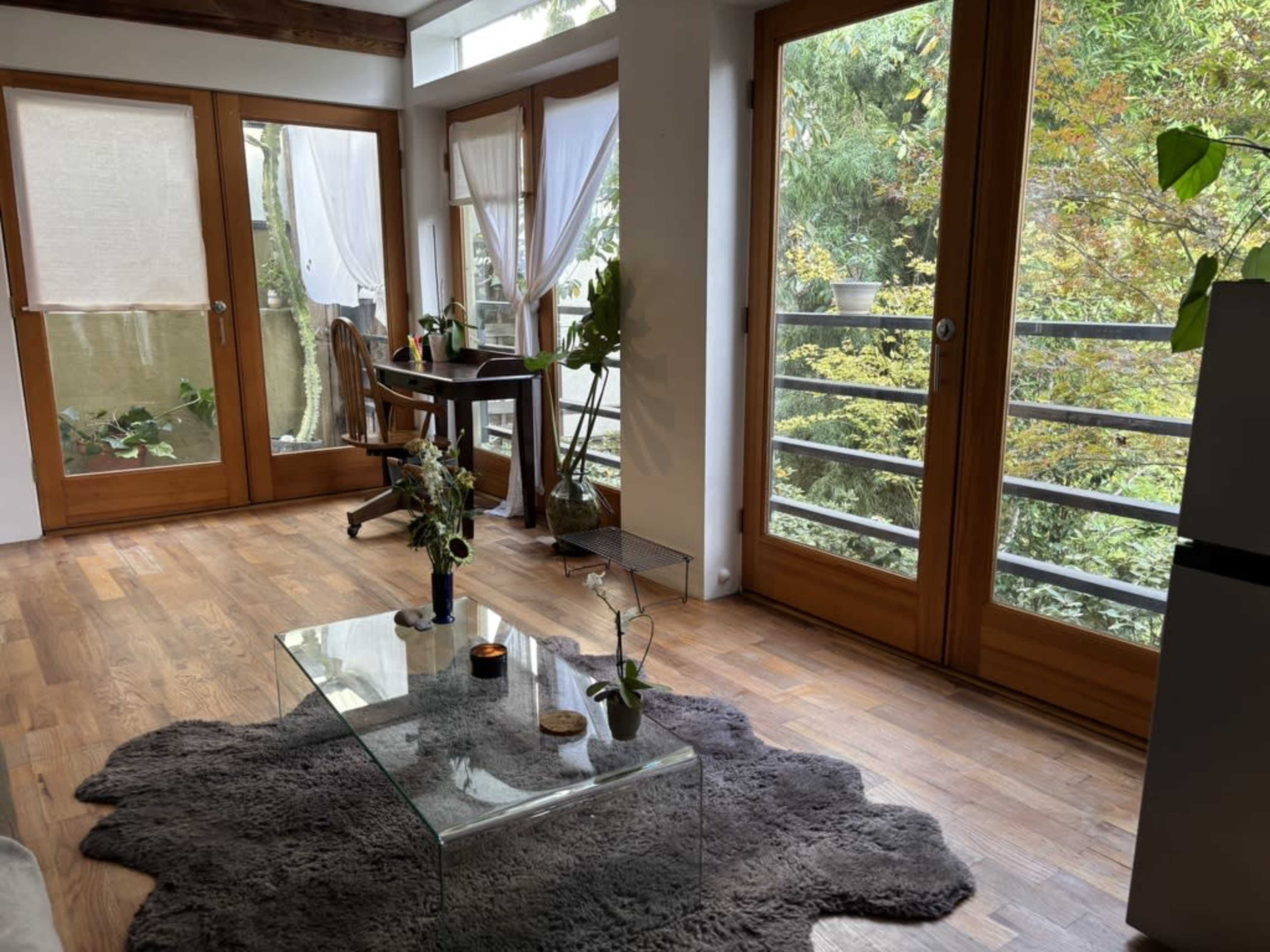 The image shows a bright living room with large windows, a glass coffee table, and a wooden table with chairs, overlooking a green outdoor area.
