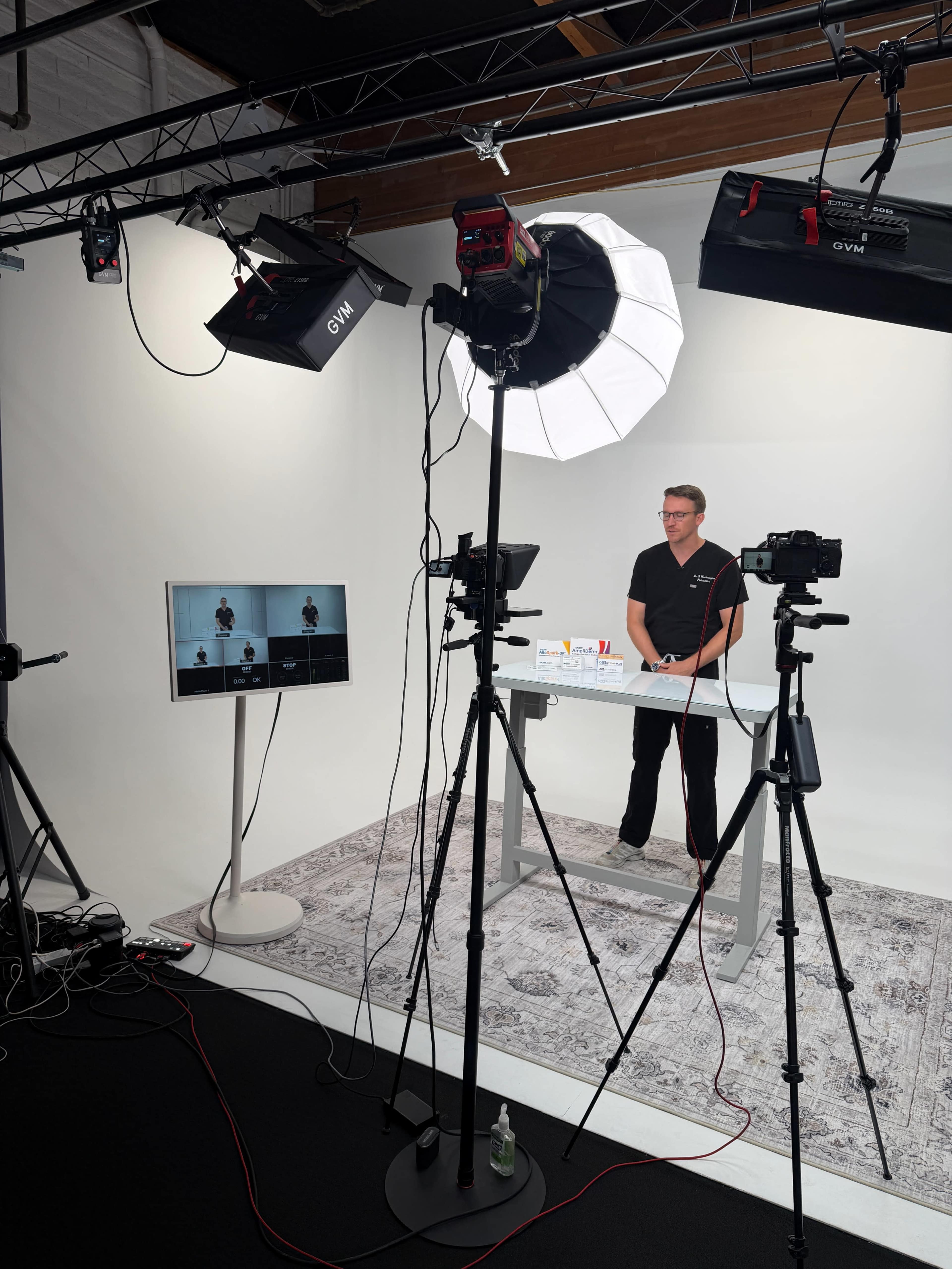 A studio setup features a table with a presenter standing behind it, surrounded by cameras, lighting equipment, and a monitor displaying the recording.