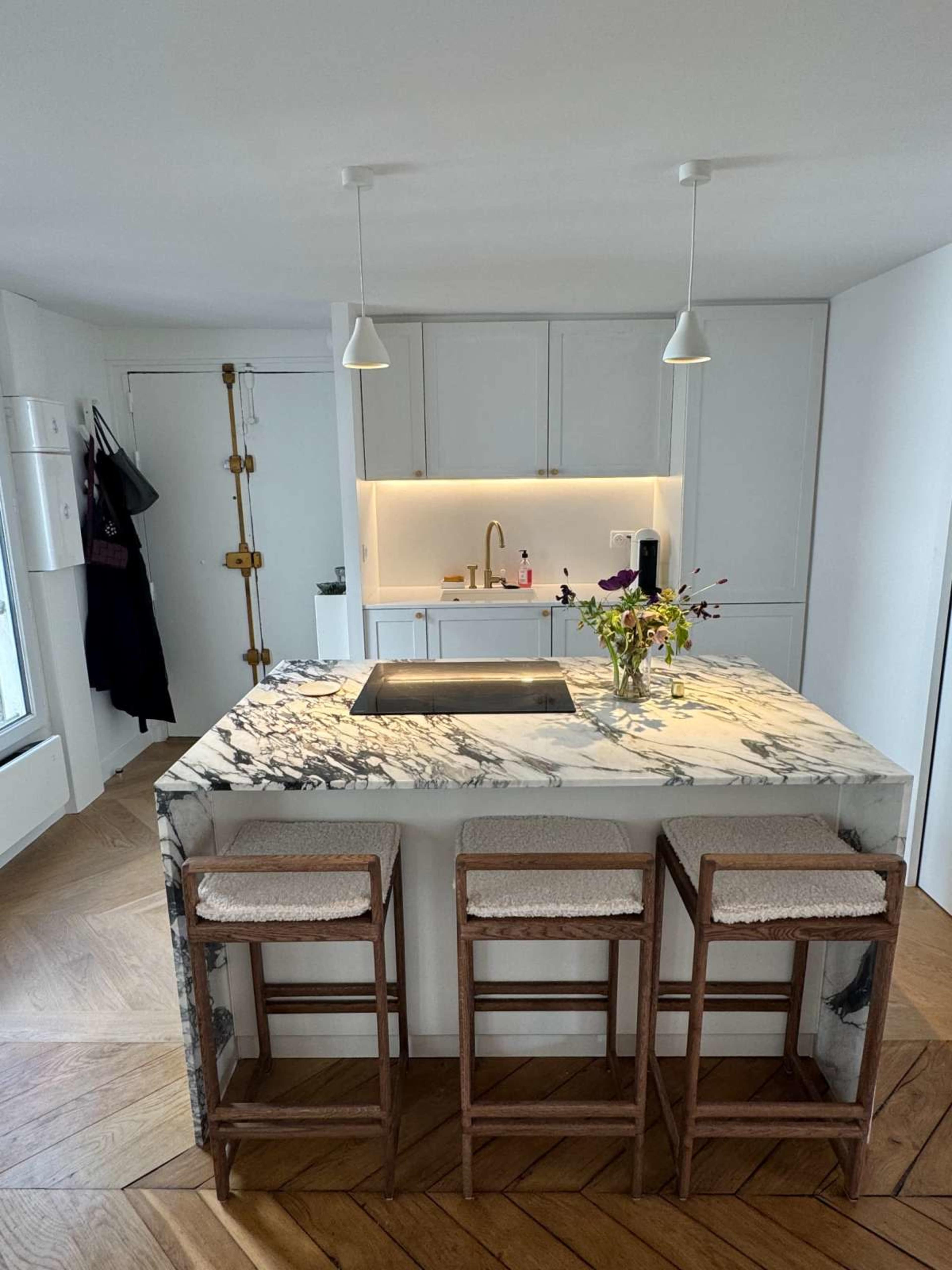 The image shows a modern kitchen with a central marble island, three wooden stools, and a minimalist design featuring white cabinetry and warm wooden flooring.