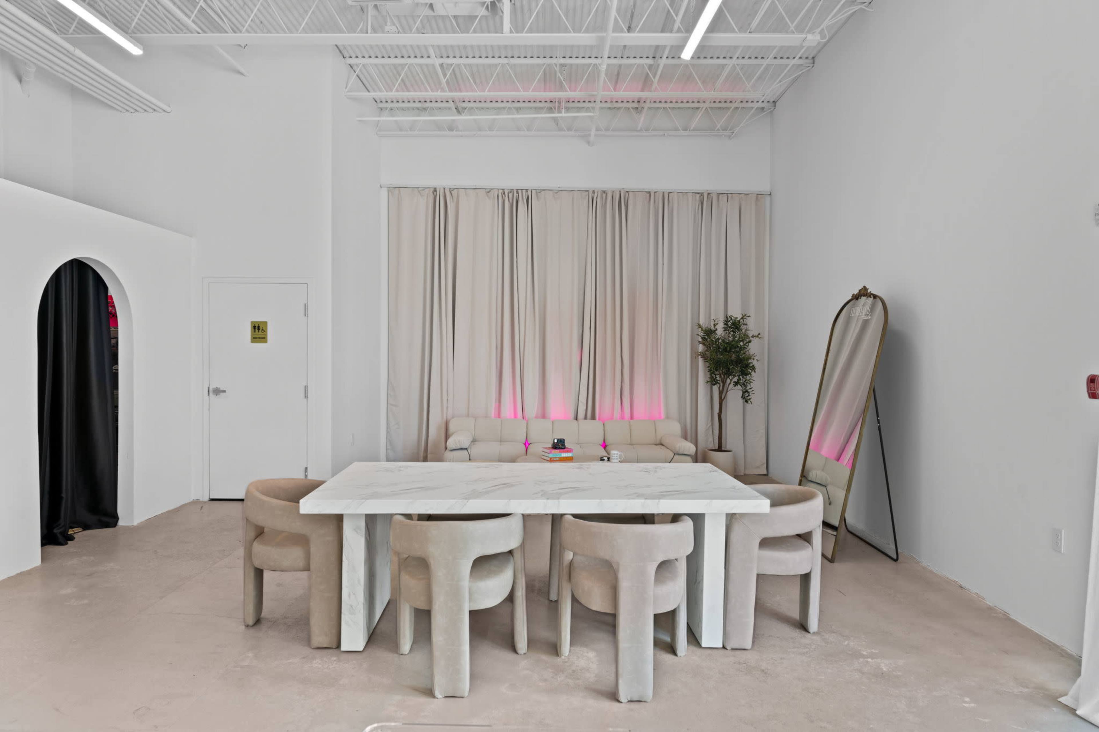 A spacious room features a large white table surrounded by beige upholstered chairs, with a light curtain backdrop and a large mirror against one wall.