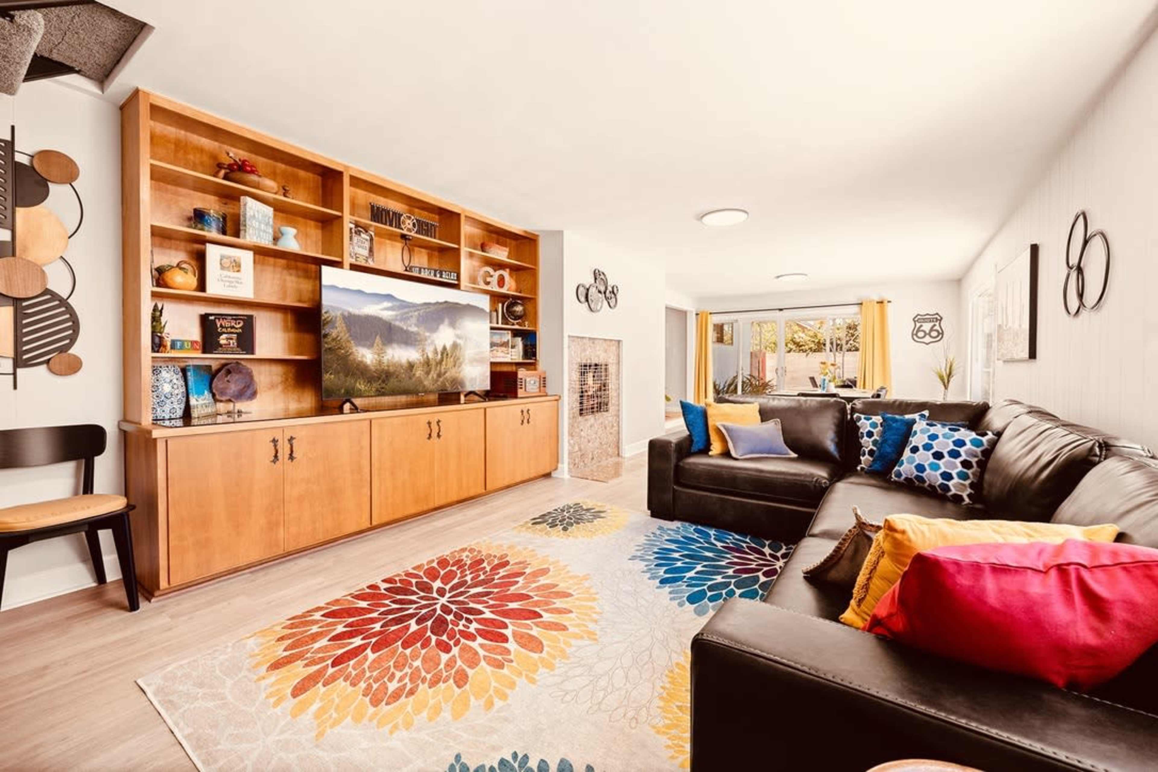 A living room featuring a large wall-mounted TV, a wooden entertainment unit, a sectional couch with colorful cushions, and a patterned area rug with floral designs.