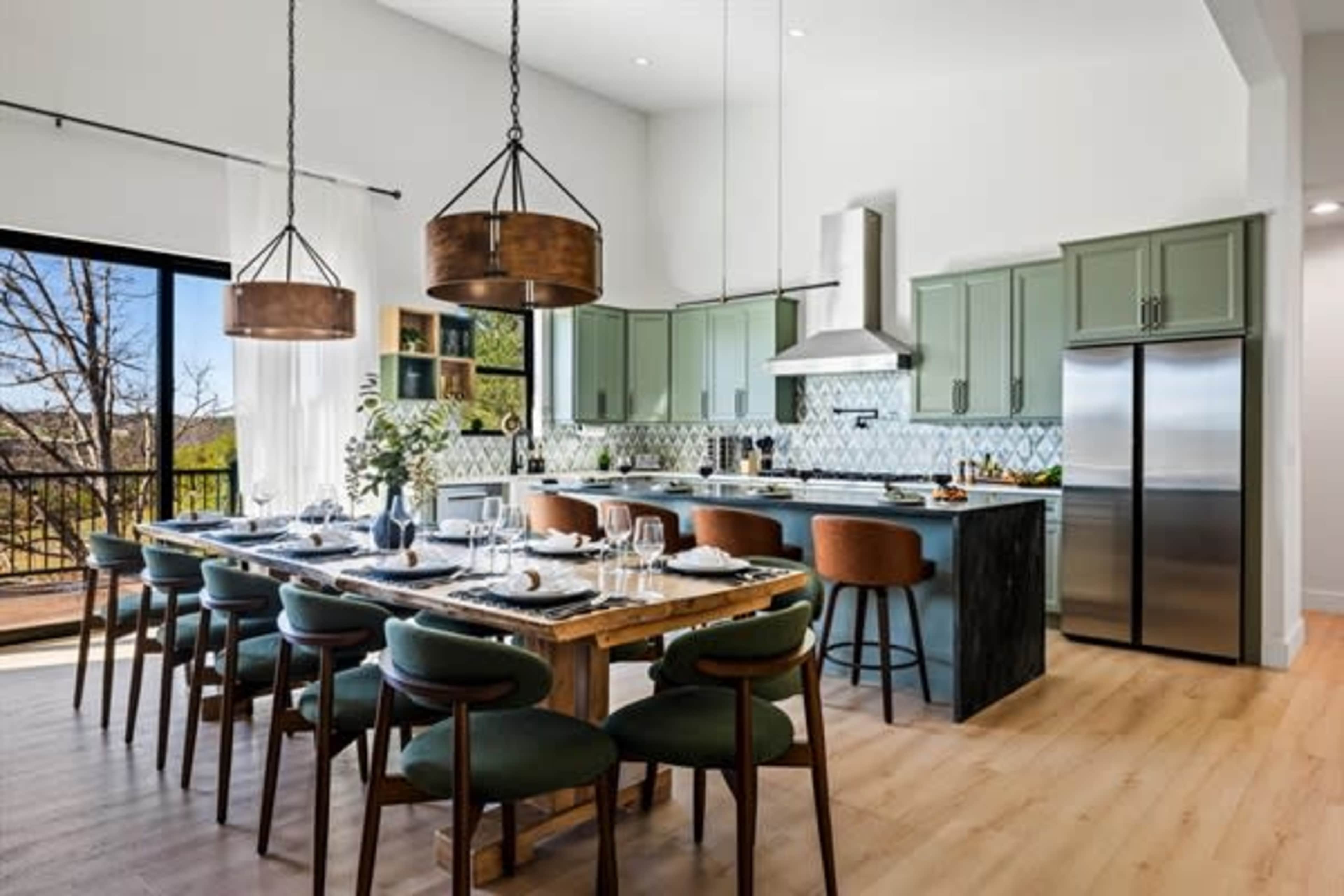 A spacious modern kitchen features a large wooden dining table surrounded by chairs, with pendant lighting above and green cabinets lining the walls.