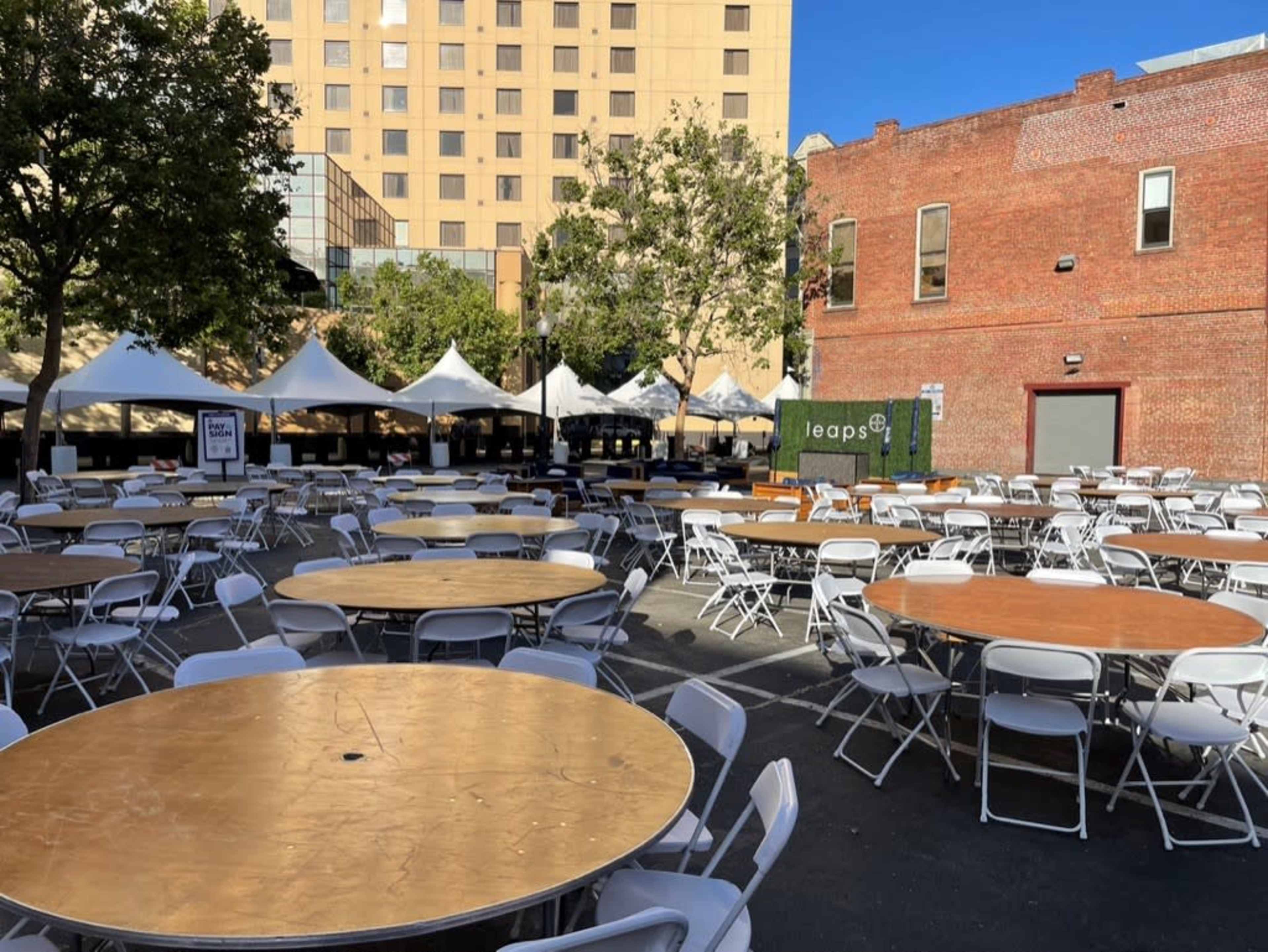 A spacious outdoor area filled with circular tables and white chairs, surrounded by tents and buildings.