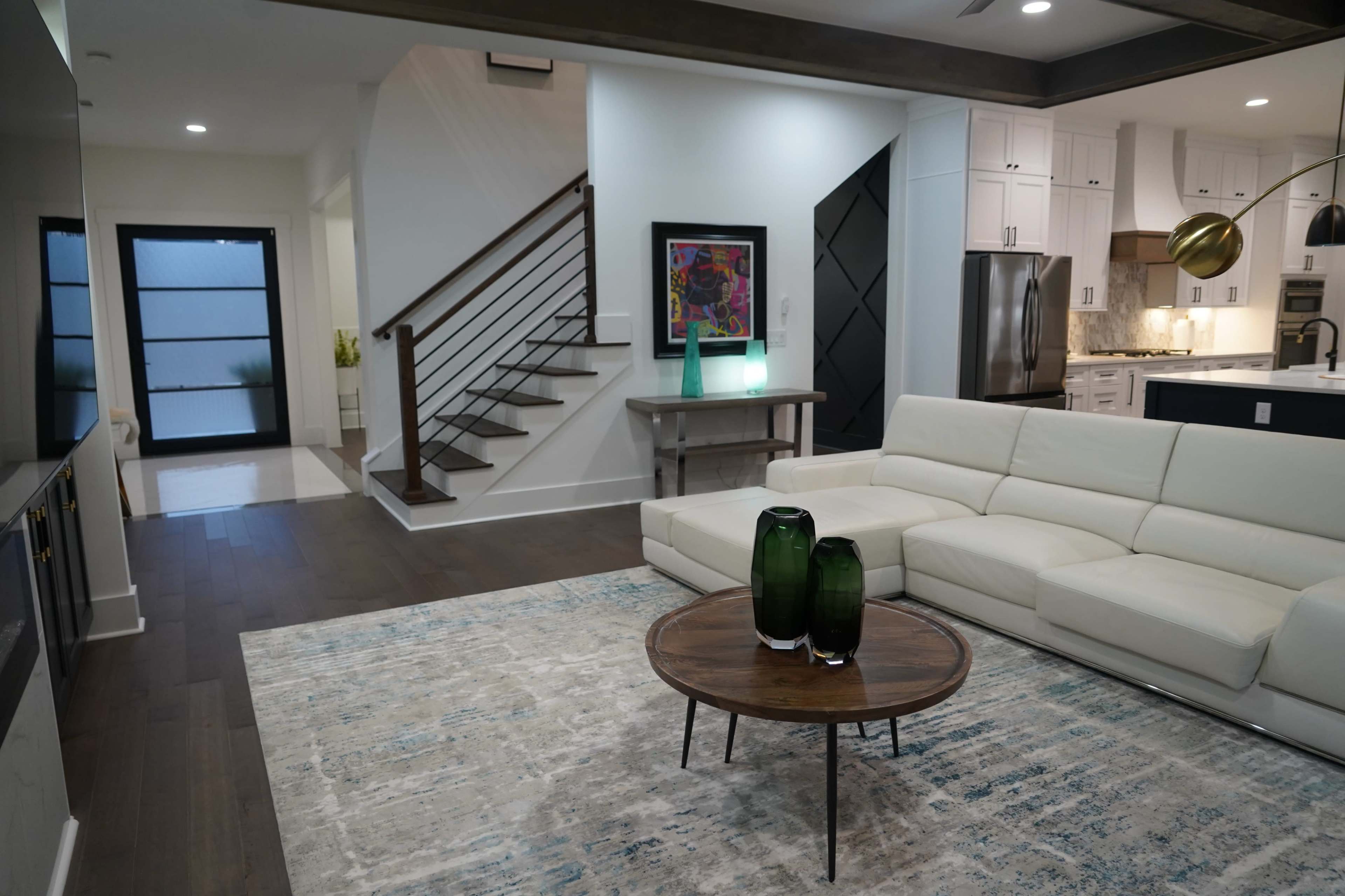 A modern living room features a white sectional sofa, a round wooden coffee table with green vases, and a staircase leading to the upper floor.