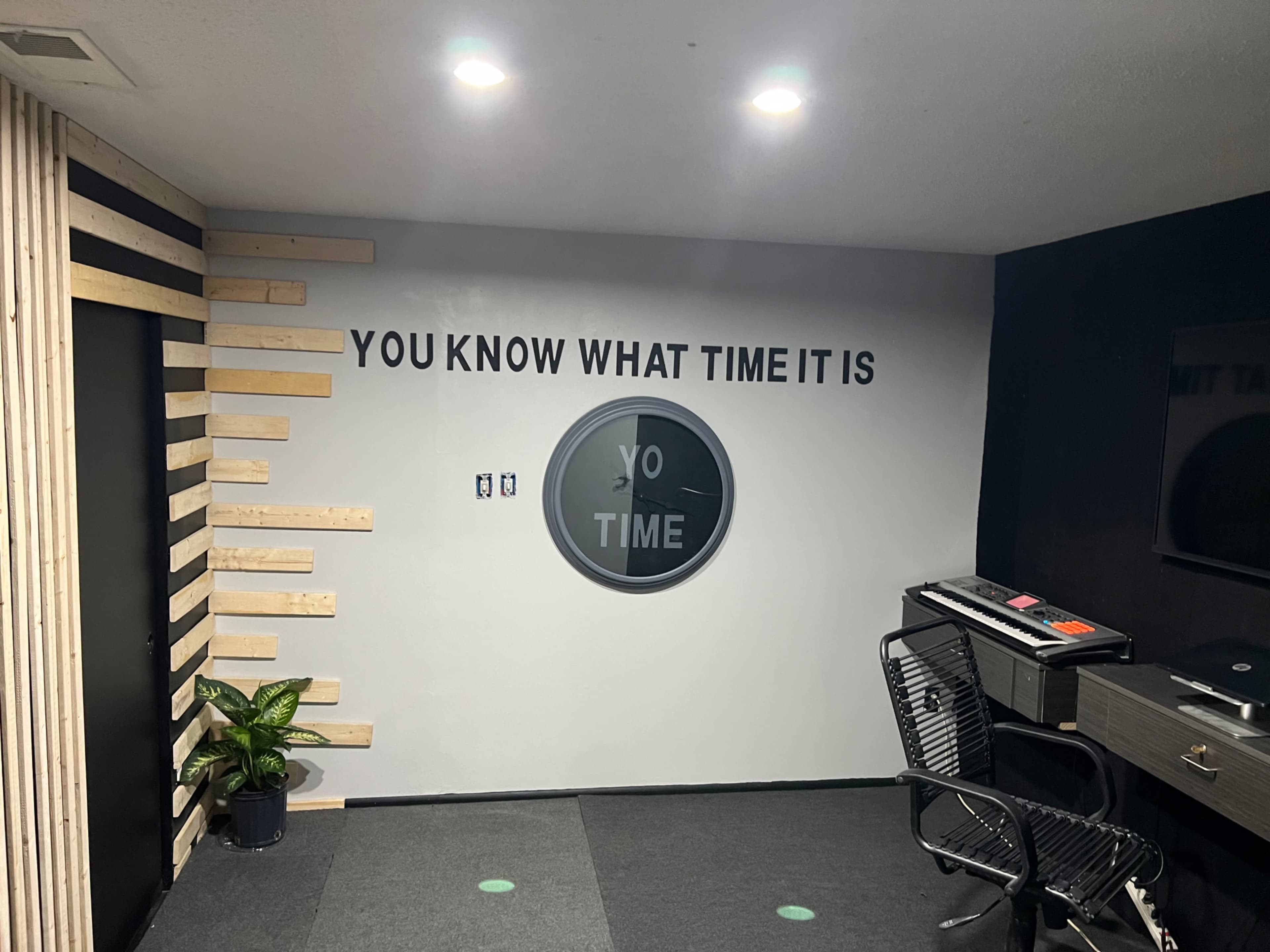 An interior wall featuring a circular clock and the text "YOU KNOW WHAT TIME IT IS" alongside a small plant and a desk area.
