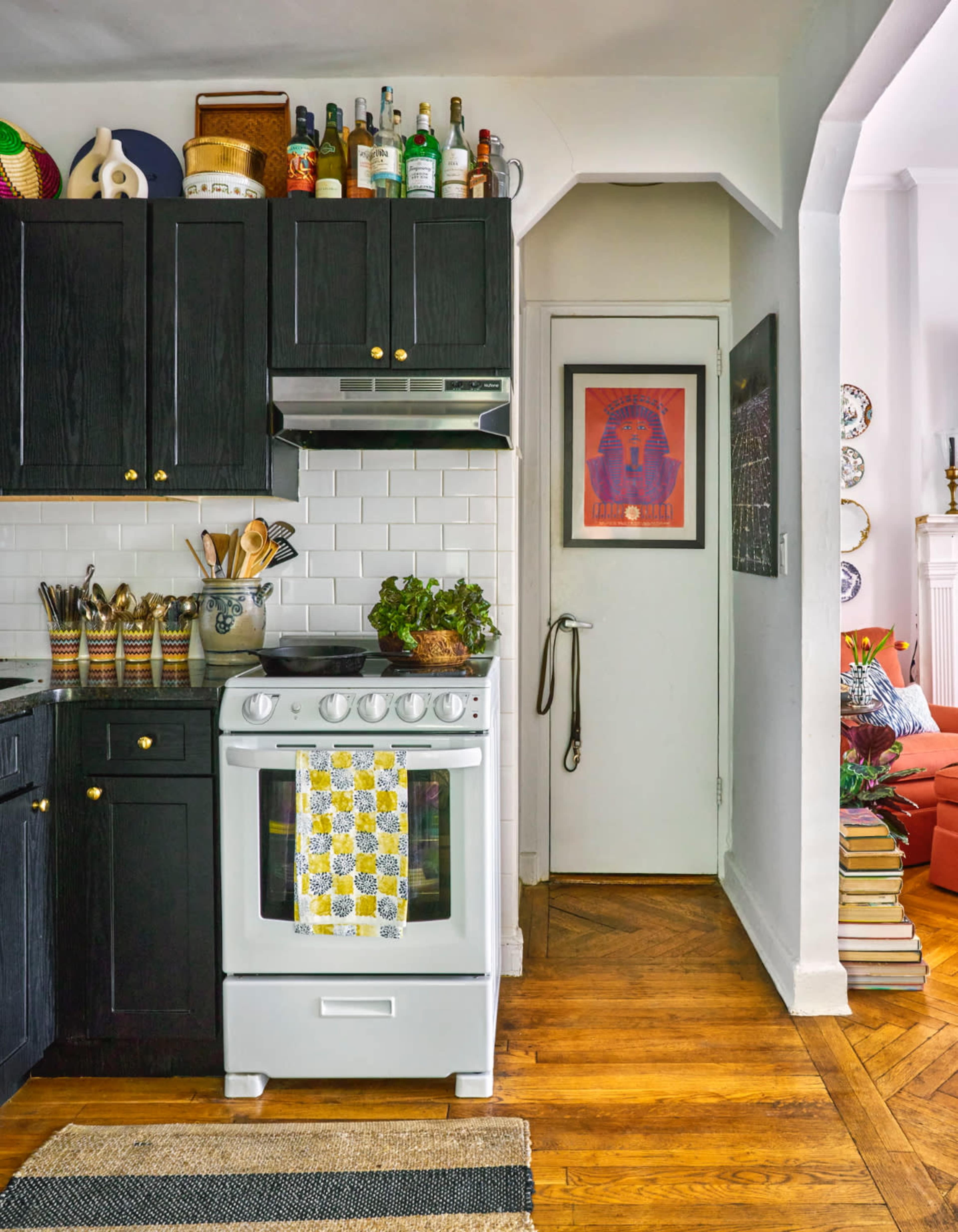 A modern kitchen features dark cabinets, a white stove, shelves lined with bottles, and an adjacent doorway leading to another room with a red sofa.