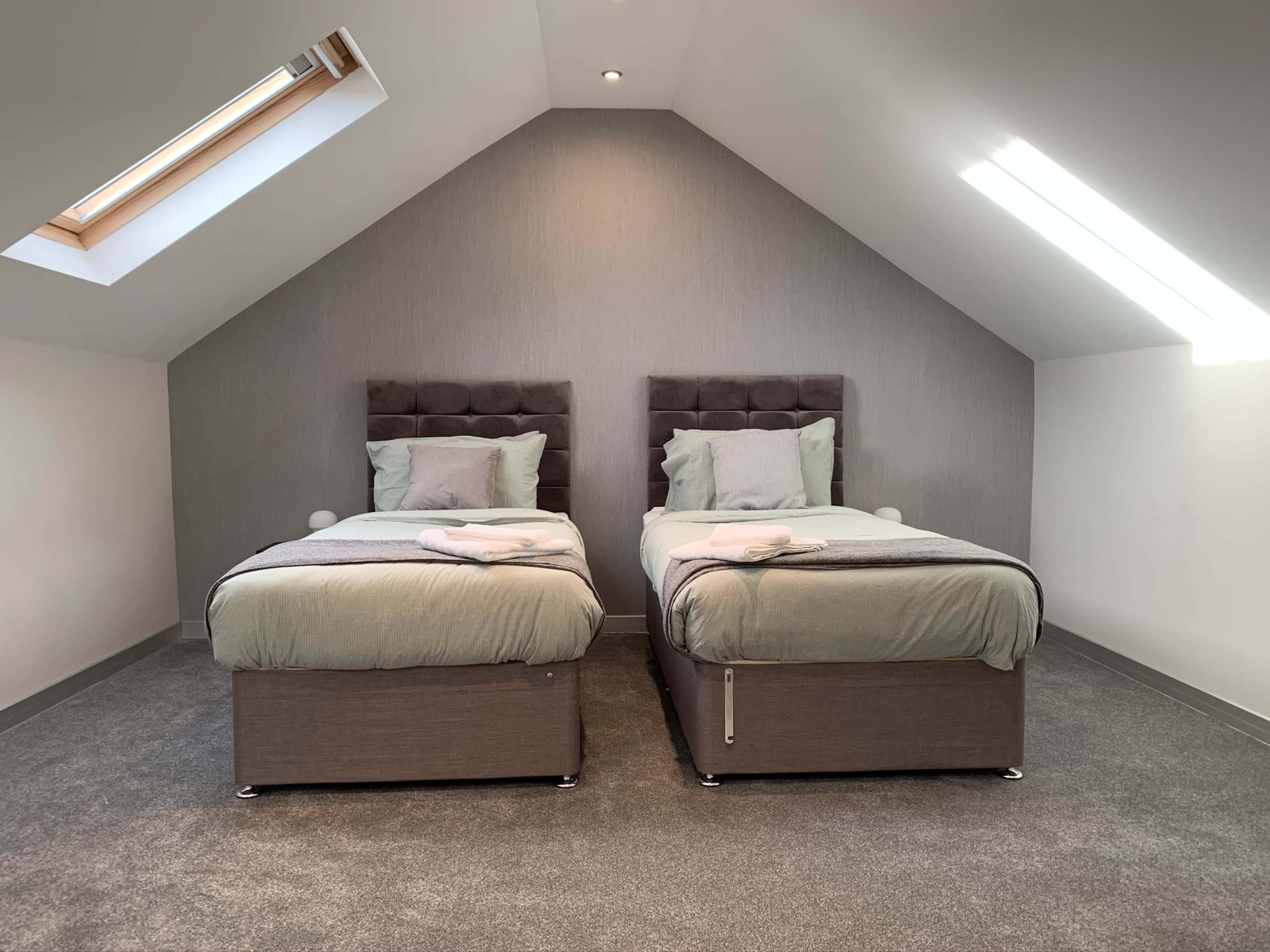 A well-lit attic bedroom with two single beds placed side by side, each featuring a gray upholstered headboard and light bedding.