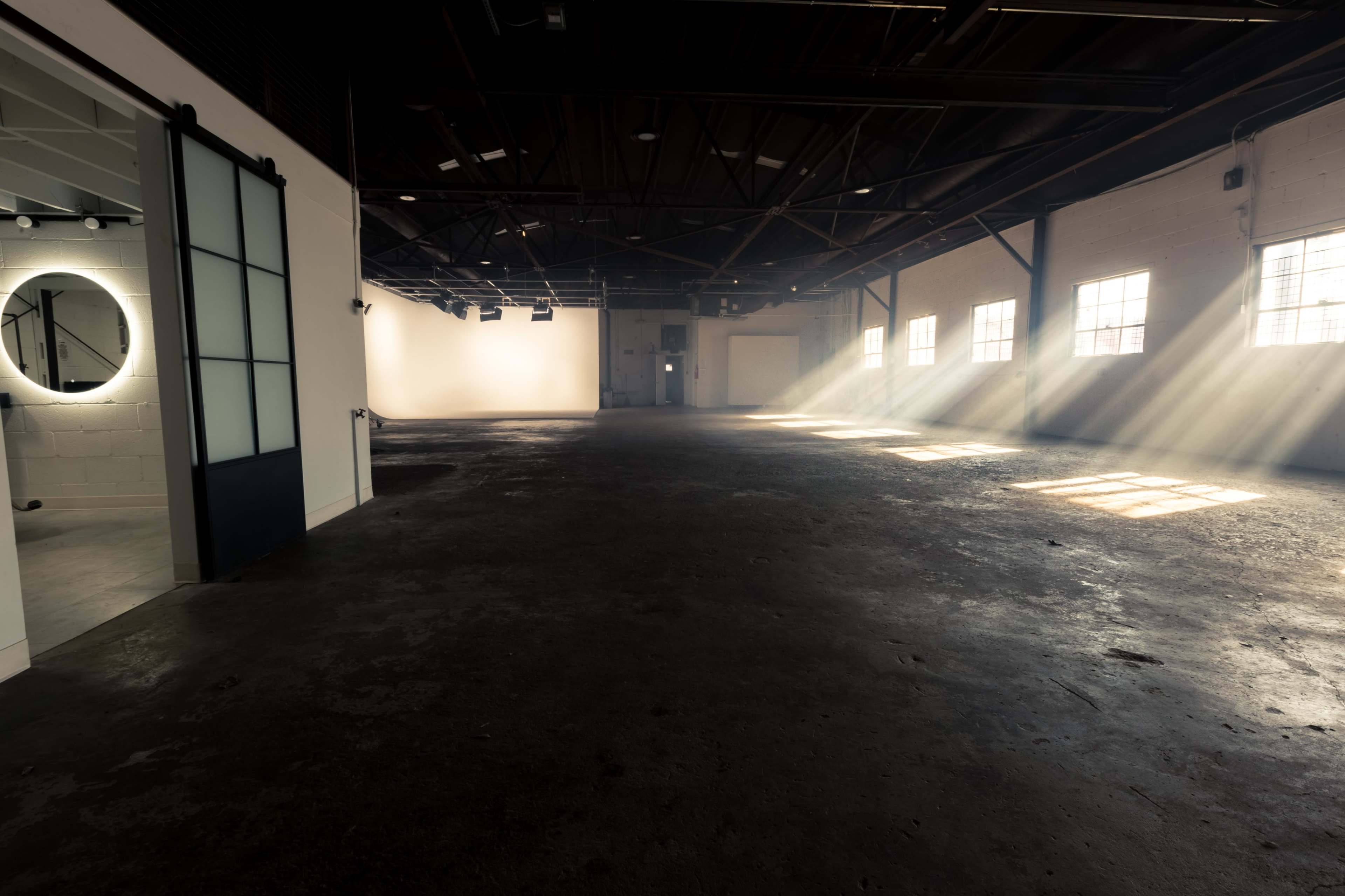 A spacious, empty industrial room with large windows allowing sunlight to stream in, creating beams of light on the floor.