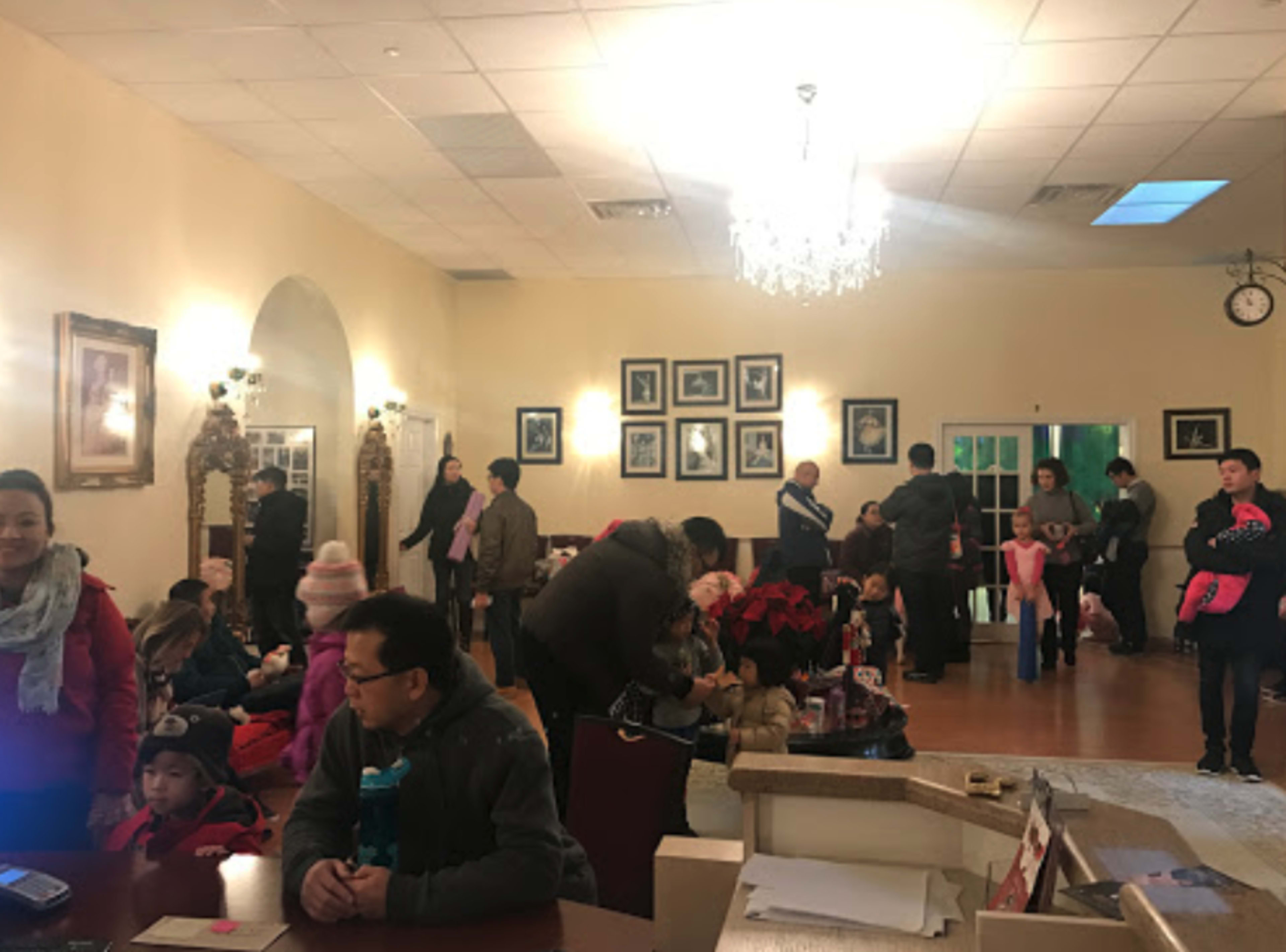 A group of people, including children, are gathered in a spacious room adorned with framed photographs and a chandelier, while some are seated and others are standing.