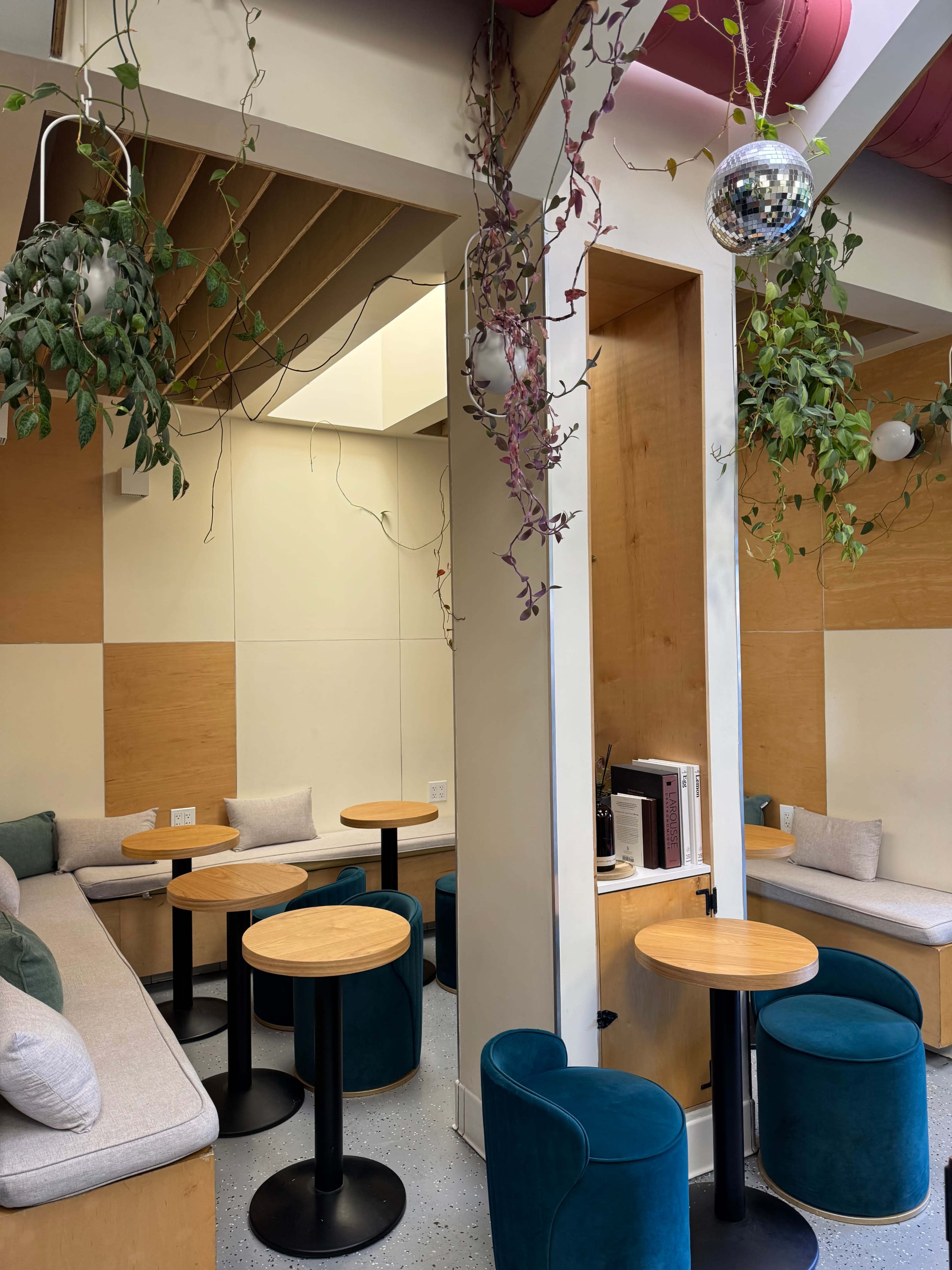 The image shows a modern, minimalistic café interior with light wooden panels, circular tables, and plush seating, adorned with hanging plants and a disco ball.