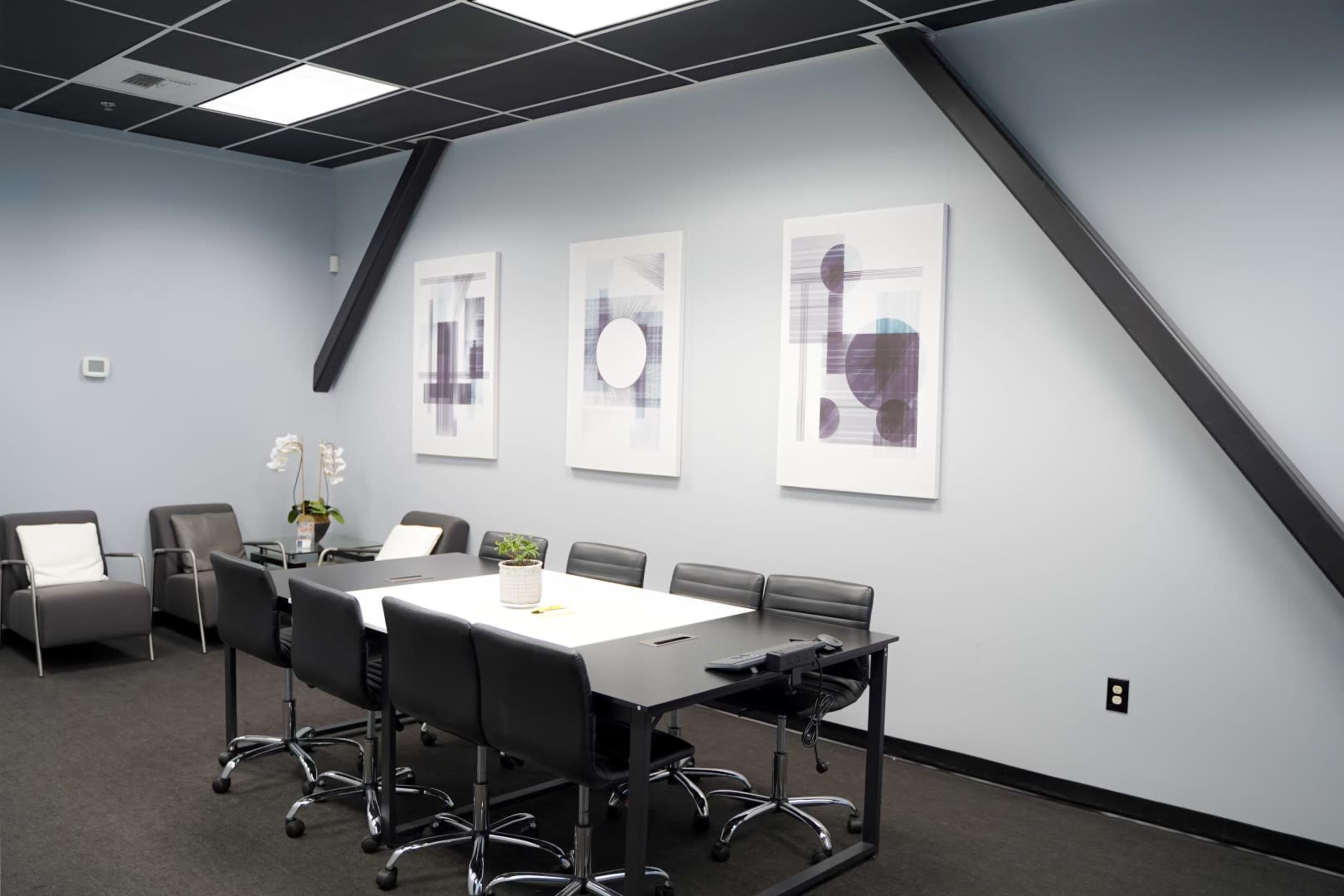 The image shows a modern conference room featuring a rectangular table surrounded by black chairs, with large framed artworks on the walls and a potted plant on the table.