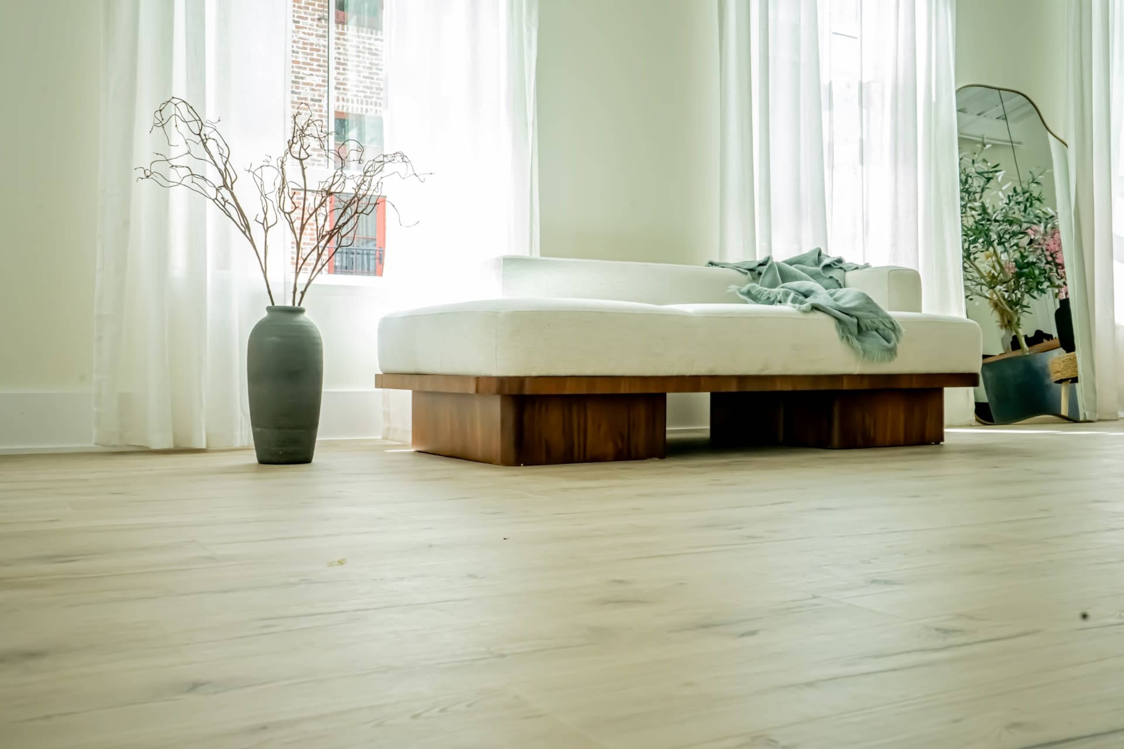 A modern living room features a white sofa on a wooden base, accompanied by a gray vase with bare branches and large windows letting in natural light.