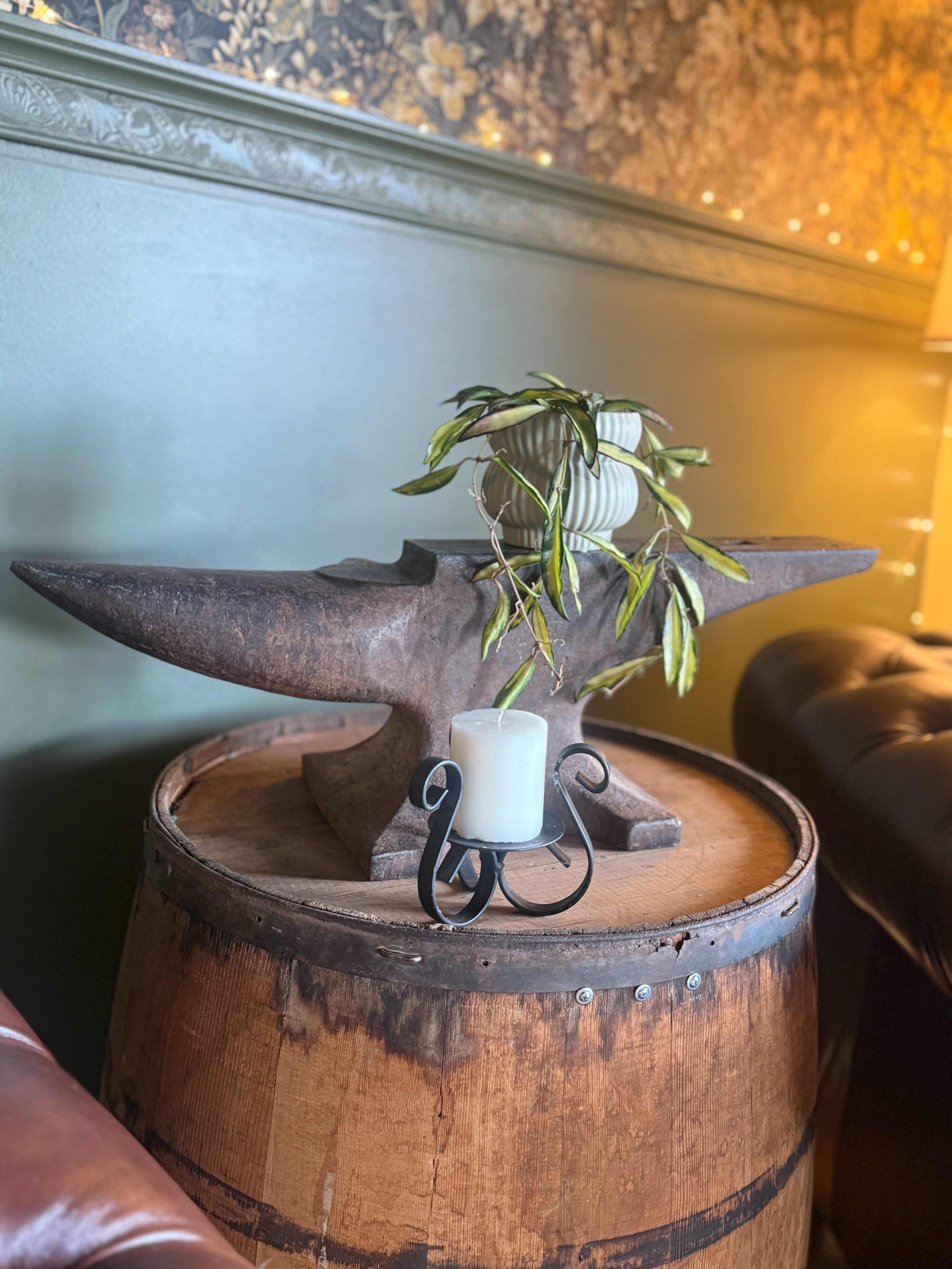 An anvil rests on top of a wooden barrel, accompanied by a small candleholder and a potted plant in a cozy indoor setting.