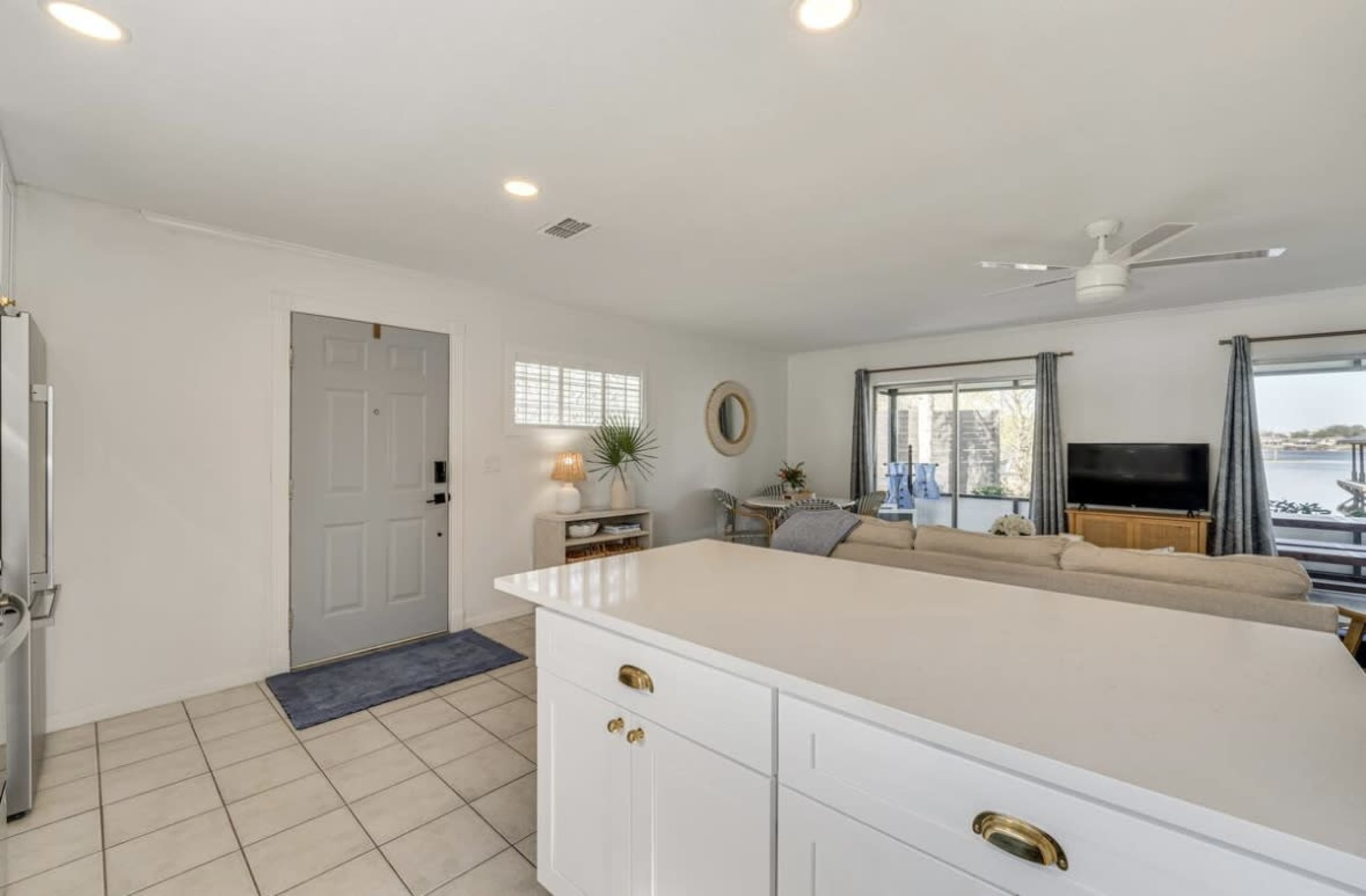 The image shows a spacious living area connected to a kitchen, featuring a front door, a large window with a view, and modern furnishings.