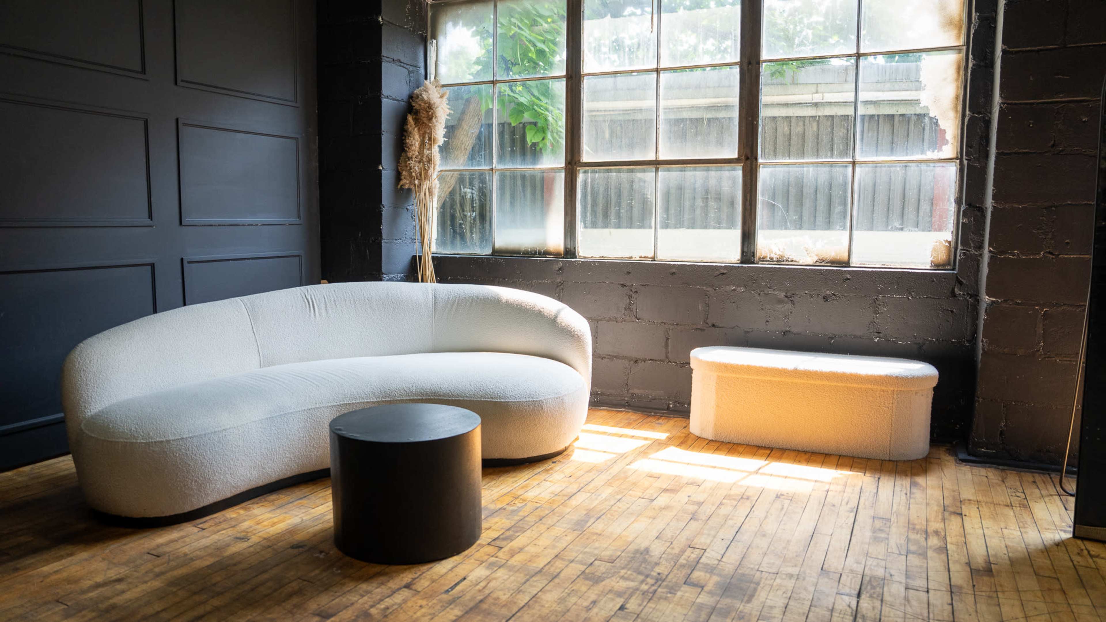 The image shows a modern, curved sofa, a round black table, and a rectangular white bench in a room with large windows and wooden flooring.