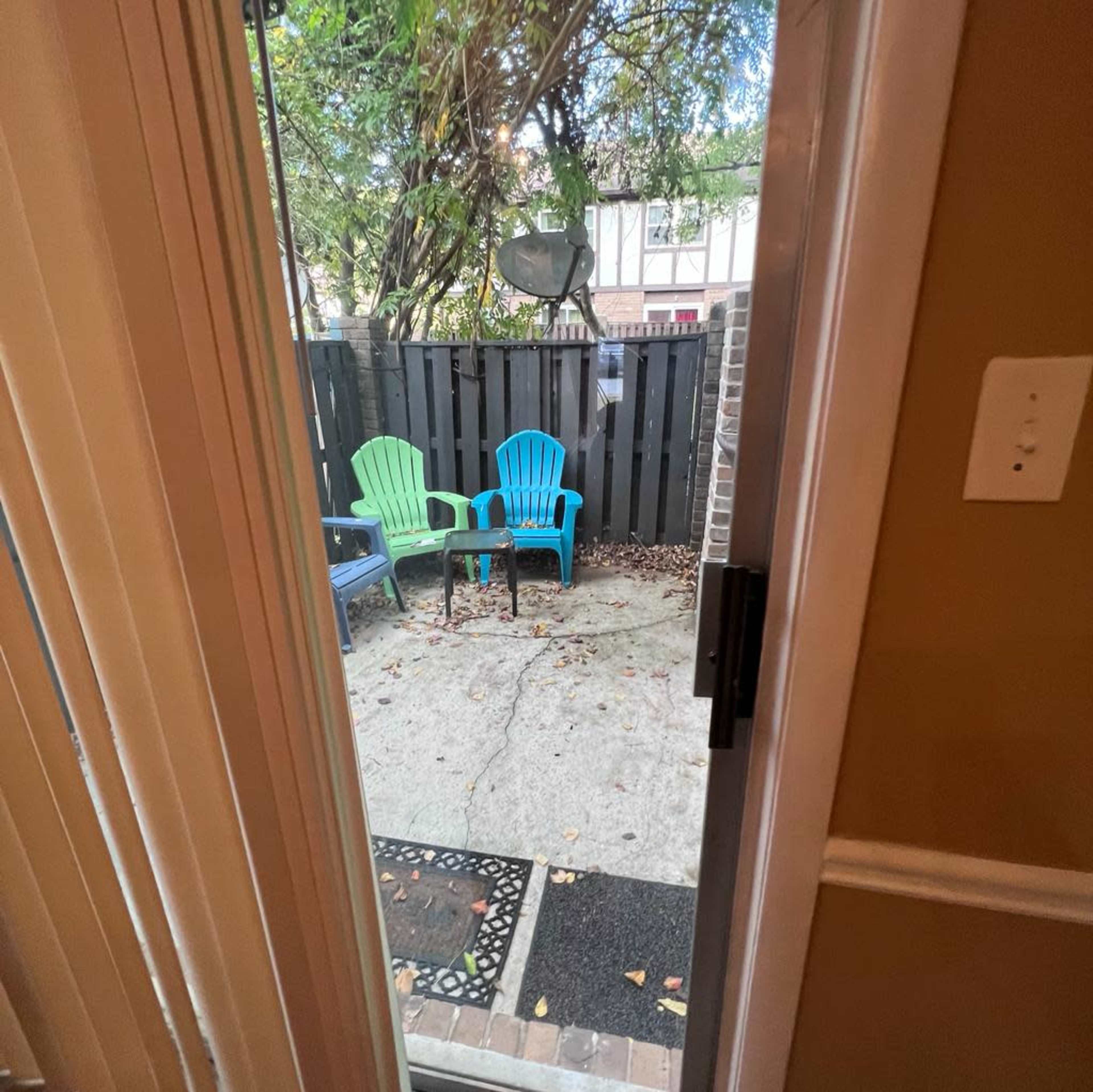 A view from inside through an open door into a small outdoor patio area with two colorful chairs and scattered leaves on the ground.
