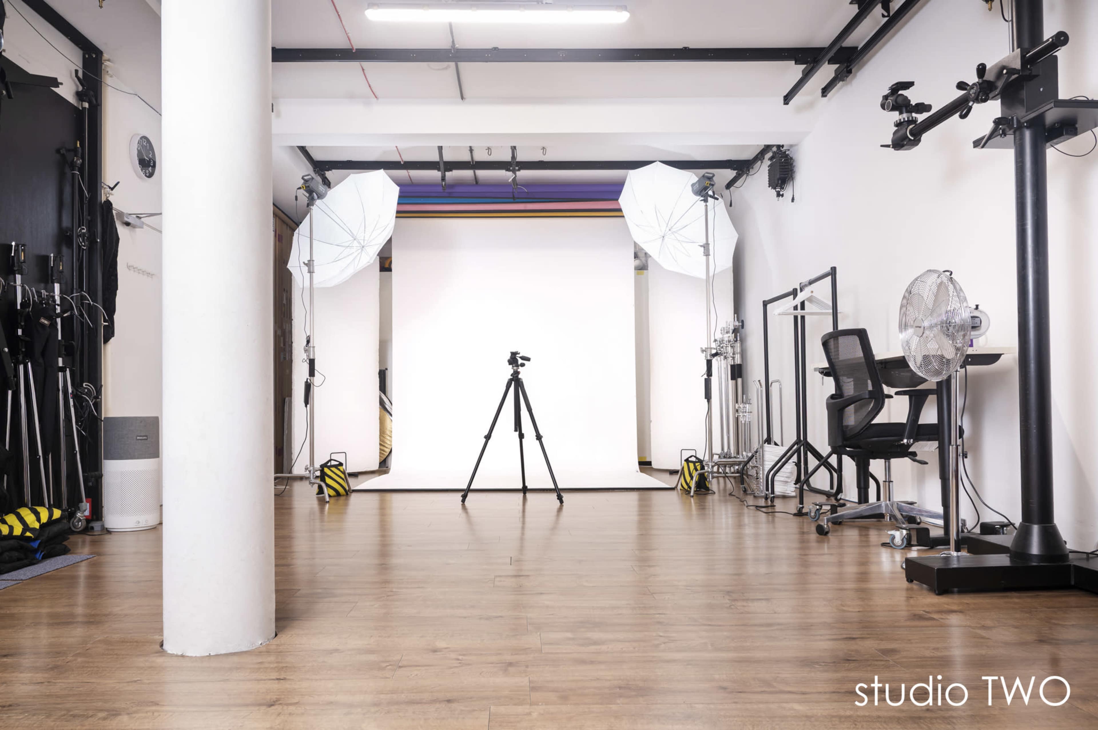 The image shows a photography studio with a white backdrop, a tripod in the center, and lighting equipment on either side.