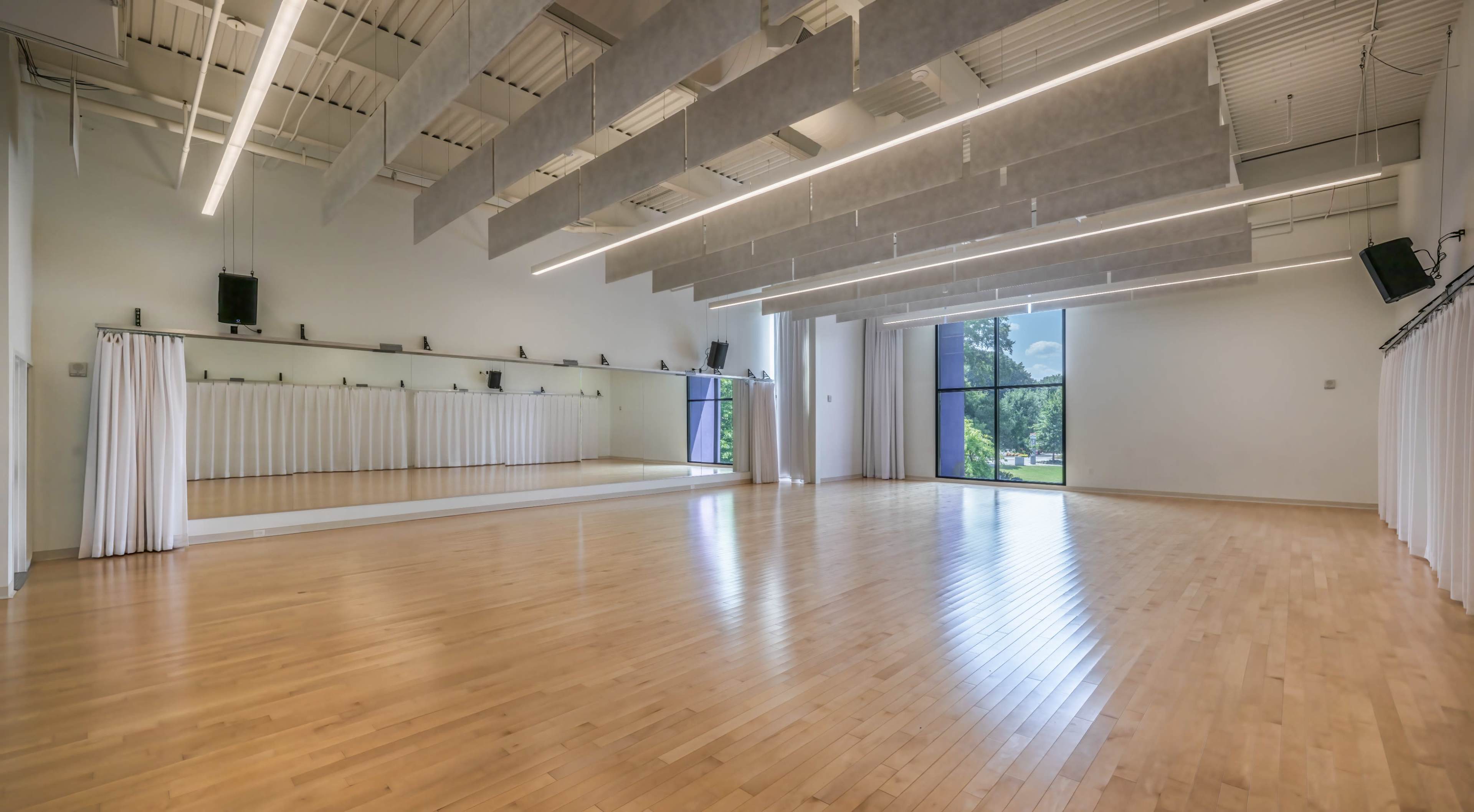 The image shows a spacious dance studio with wooden floors, large windows, and a mirrored wall.