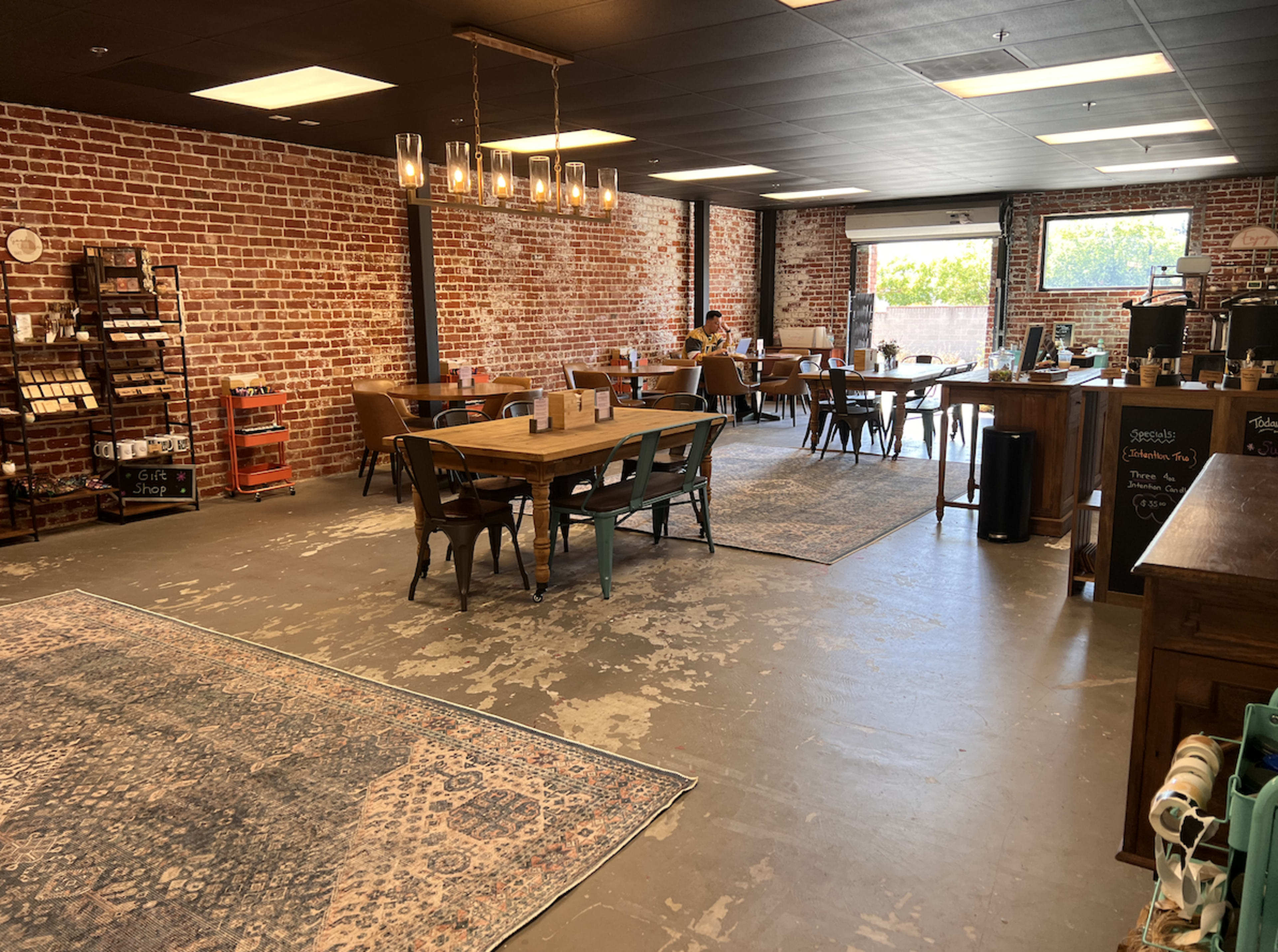 The image shows a spacious café with exposed brick walls, wooden tables, and a few chairs scattered around, featuring large windows that allow natural light to enter.