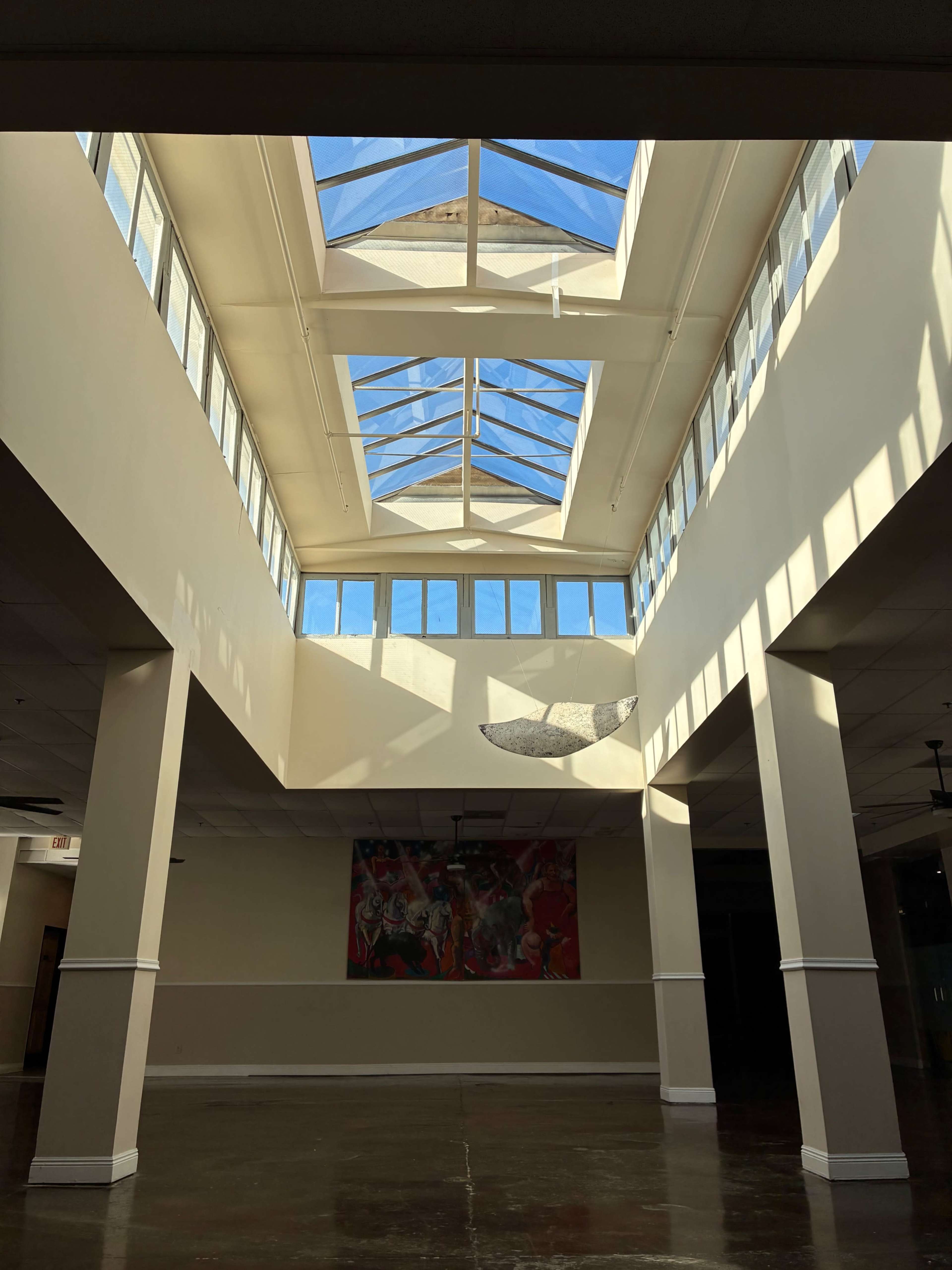 The image shows a spacious hallway with large skylights and a mural on the far wall, illuminated by natural light.