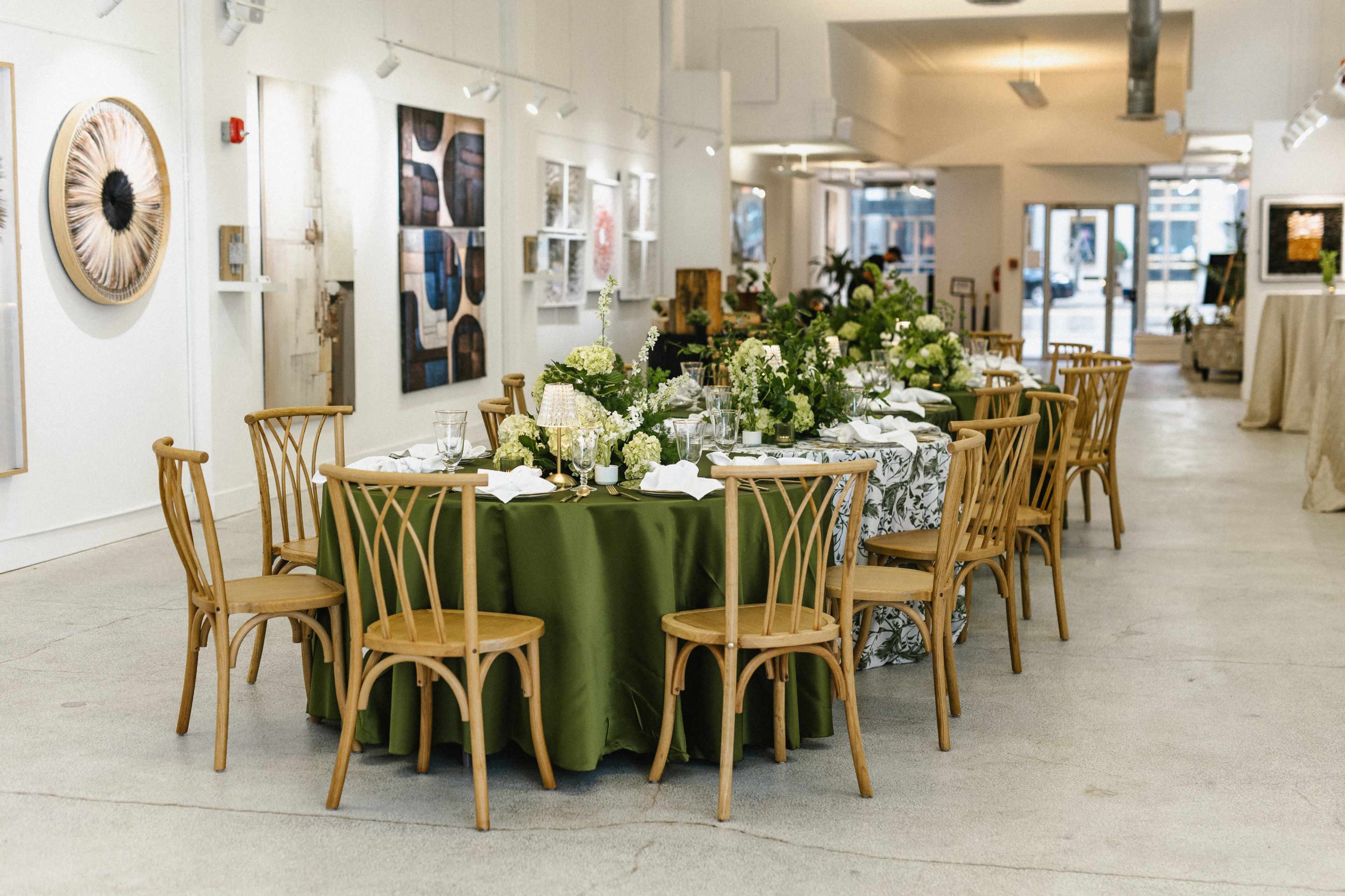 The image shows a set dining area with two tables, one covered in a green tablecloth and the other in a patterned cloth, arranged with chairs and decorative centerpieces in an art gallery.