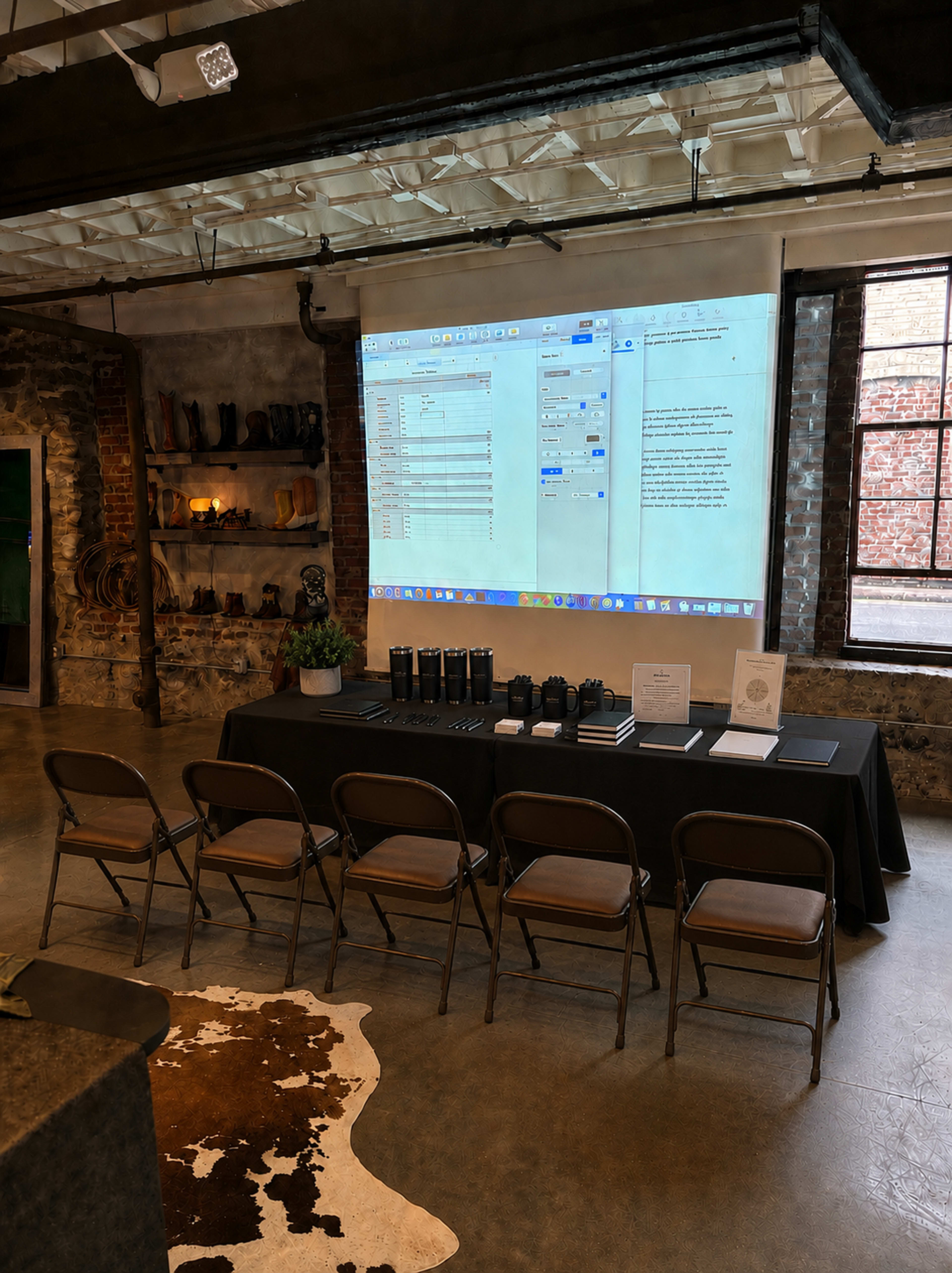 The image shows a room with a long table covered in black, featuring several items, and a large screen displaying computer files on the wall above it.