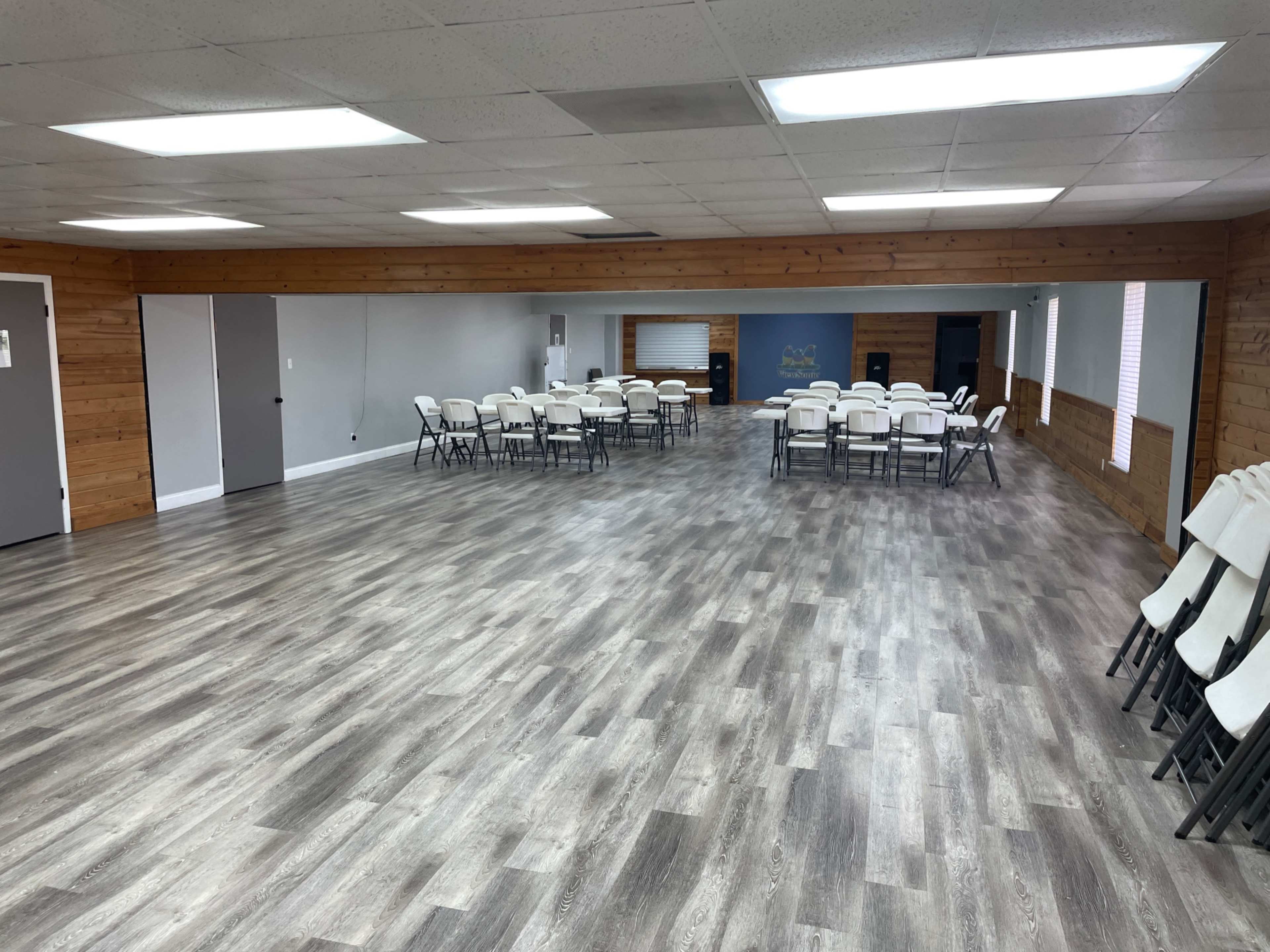 The image shows an interior room with a polished wooden floor, where several rows of folding chairs are stacked against the wall, and a number of tables are set up in the center.