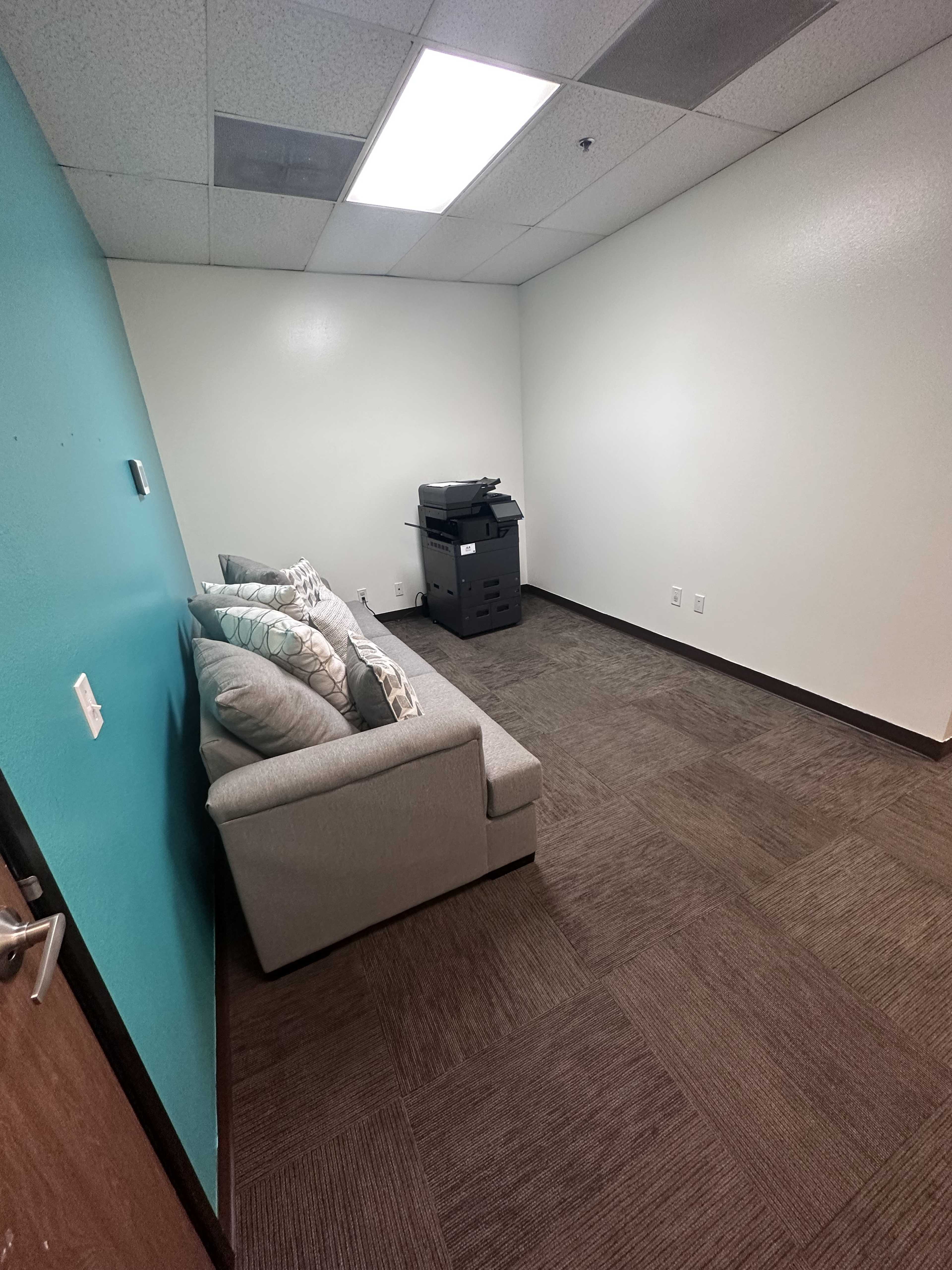 A small, empty room with a light blue wall, a gray couch with pillows, and a printer stationed in the corner.