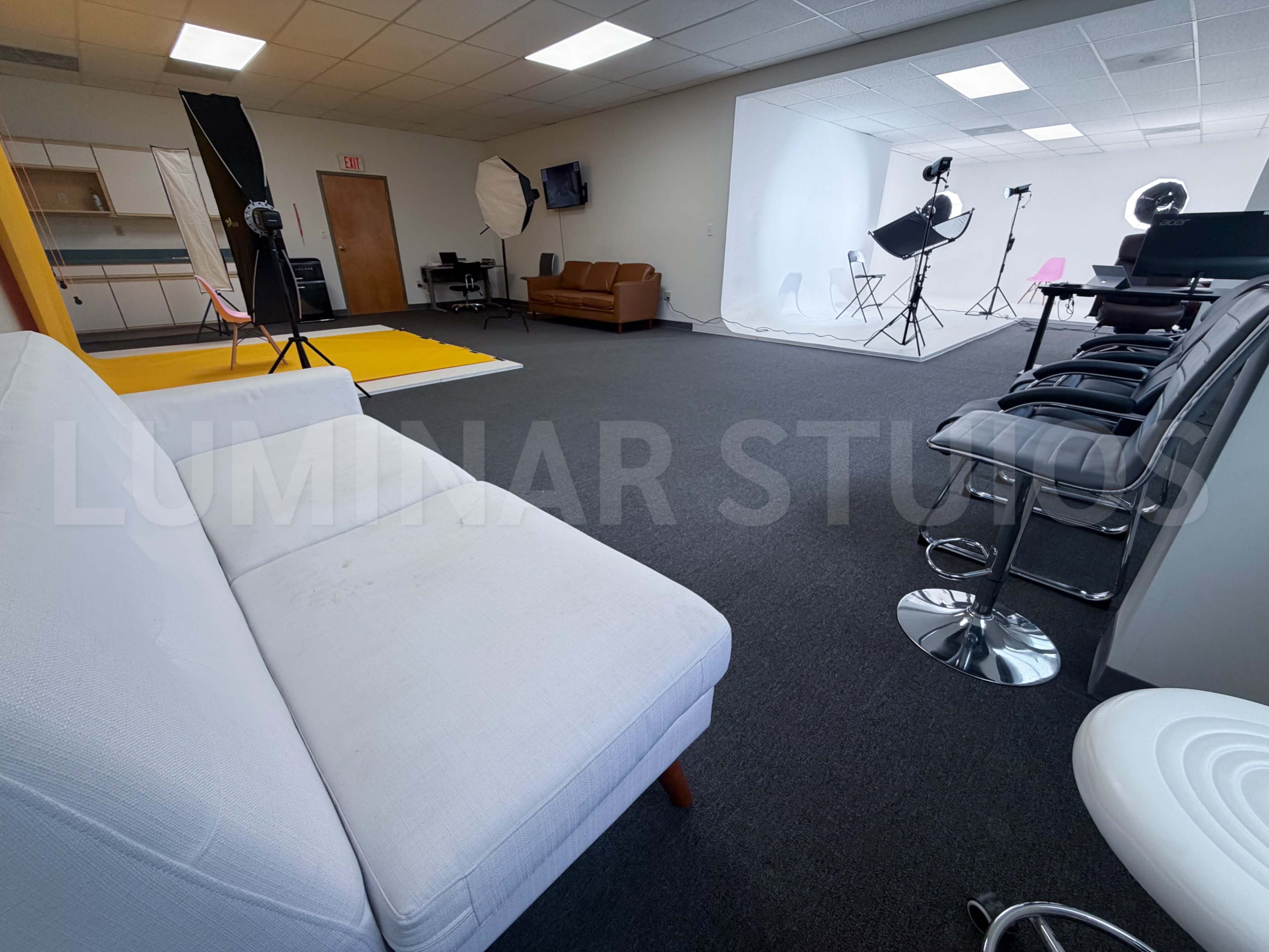 The image shows a spacious studio with a white couch, a brown leather sofa, and various photography equipment set up against a plain backdrop.