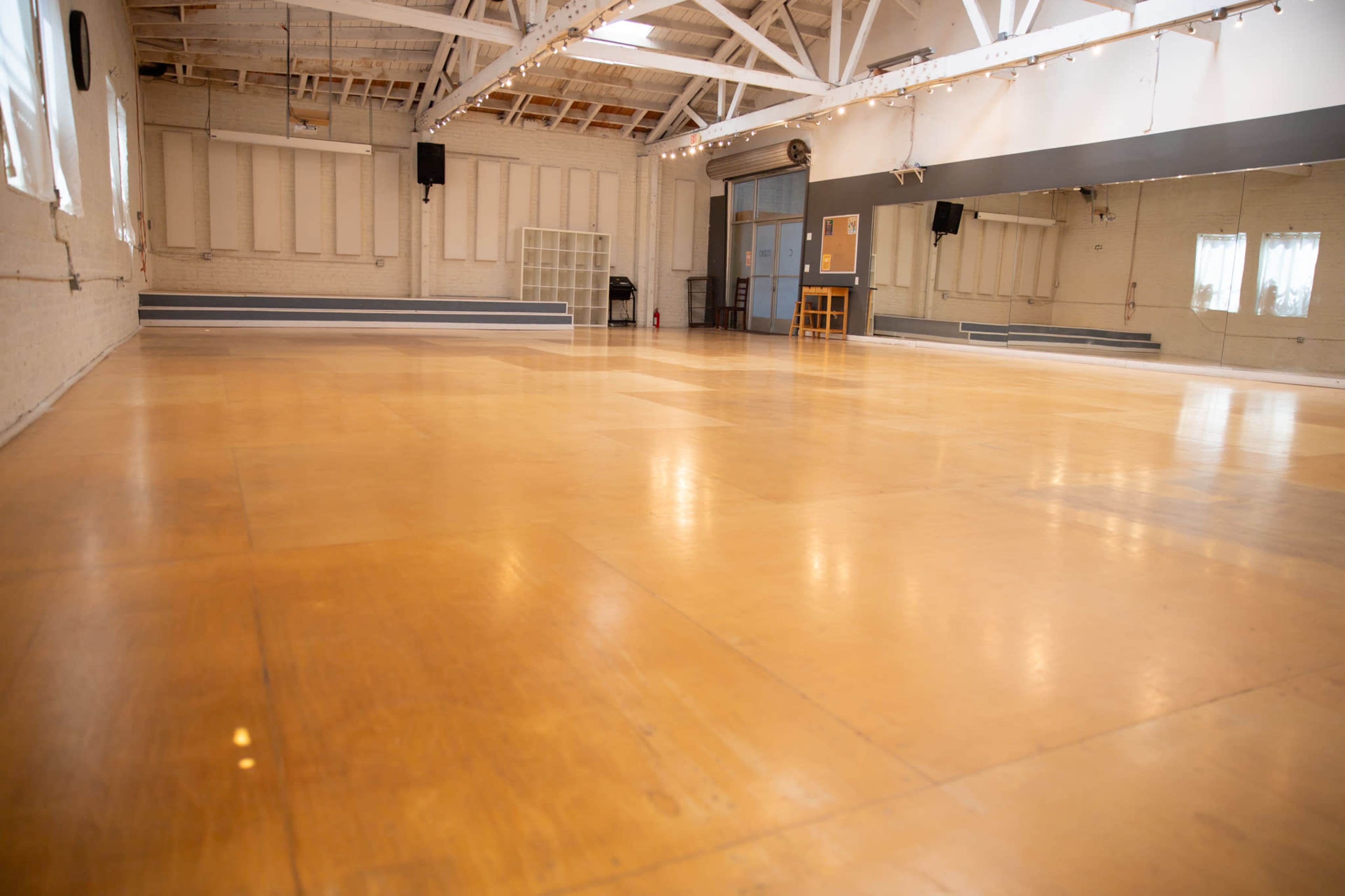The image shows an empty dance studio with wooden flooring, white walls, and large windows allowing natural light to enter.