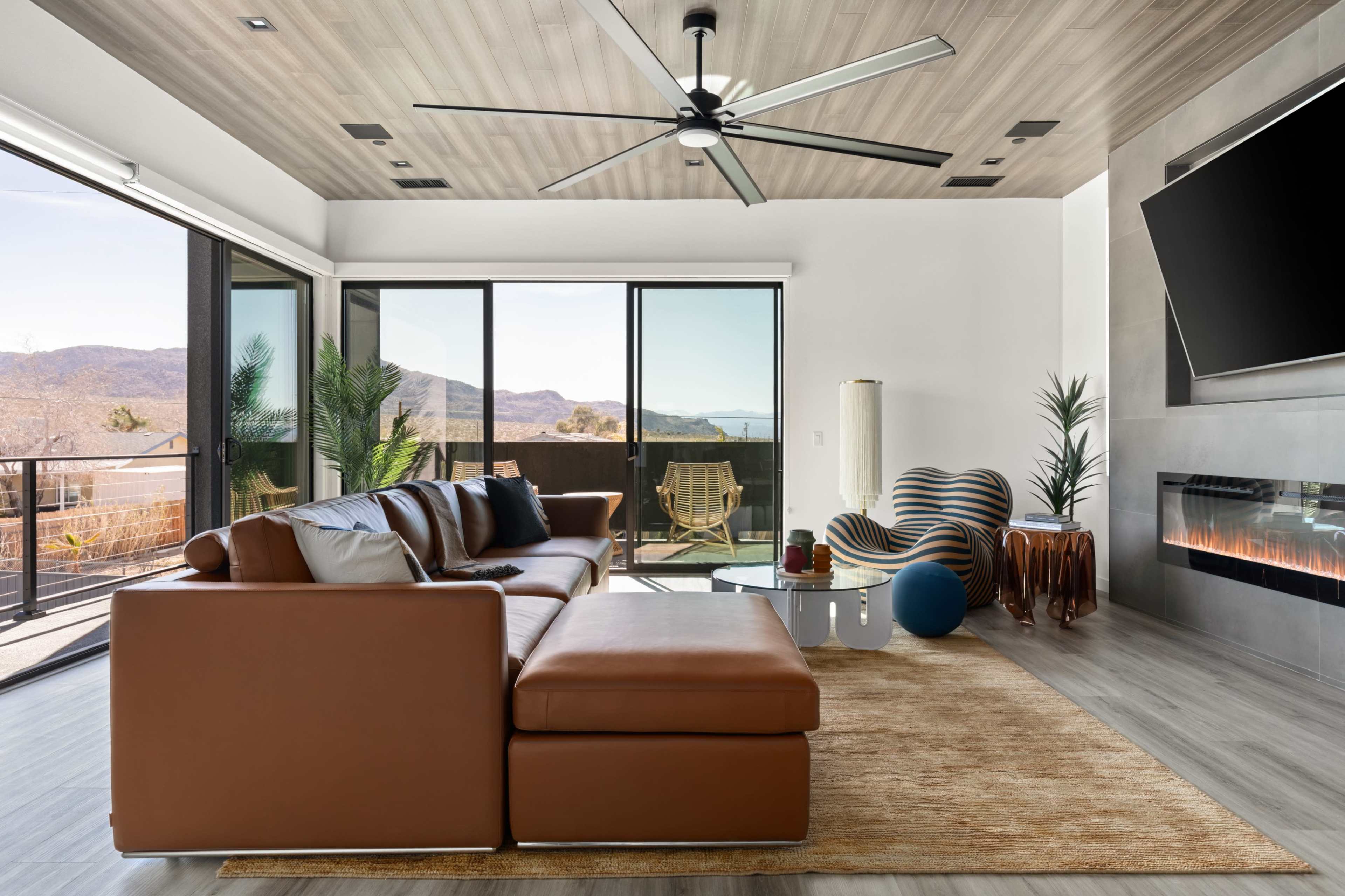 The image shows a modern living room with a brown leather sectional sofa, a glass coffee table, and large windows that overlook a mountainous landscape.