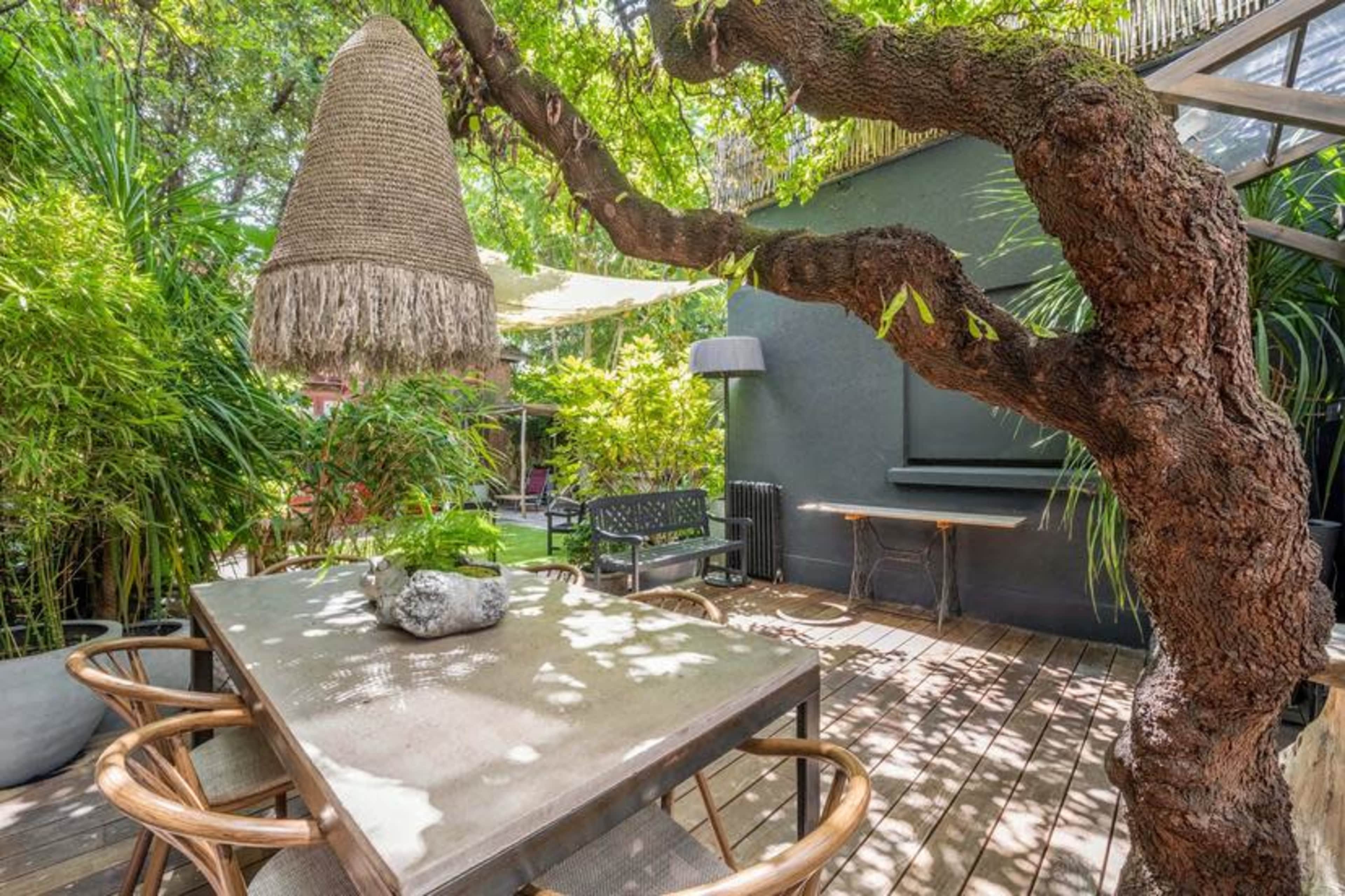 A wooden table with wicker chairs sits beneath a large tree featuring a hanging decorative light, surrounded by lush greenery.