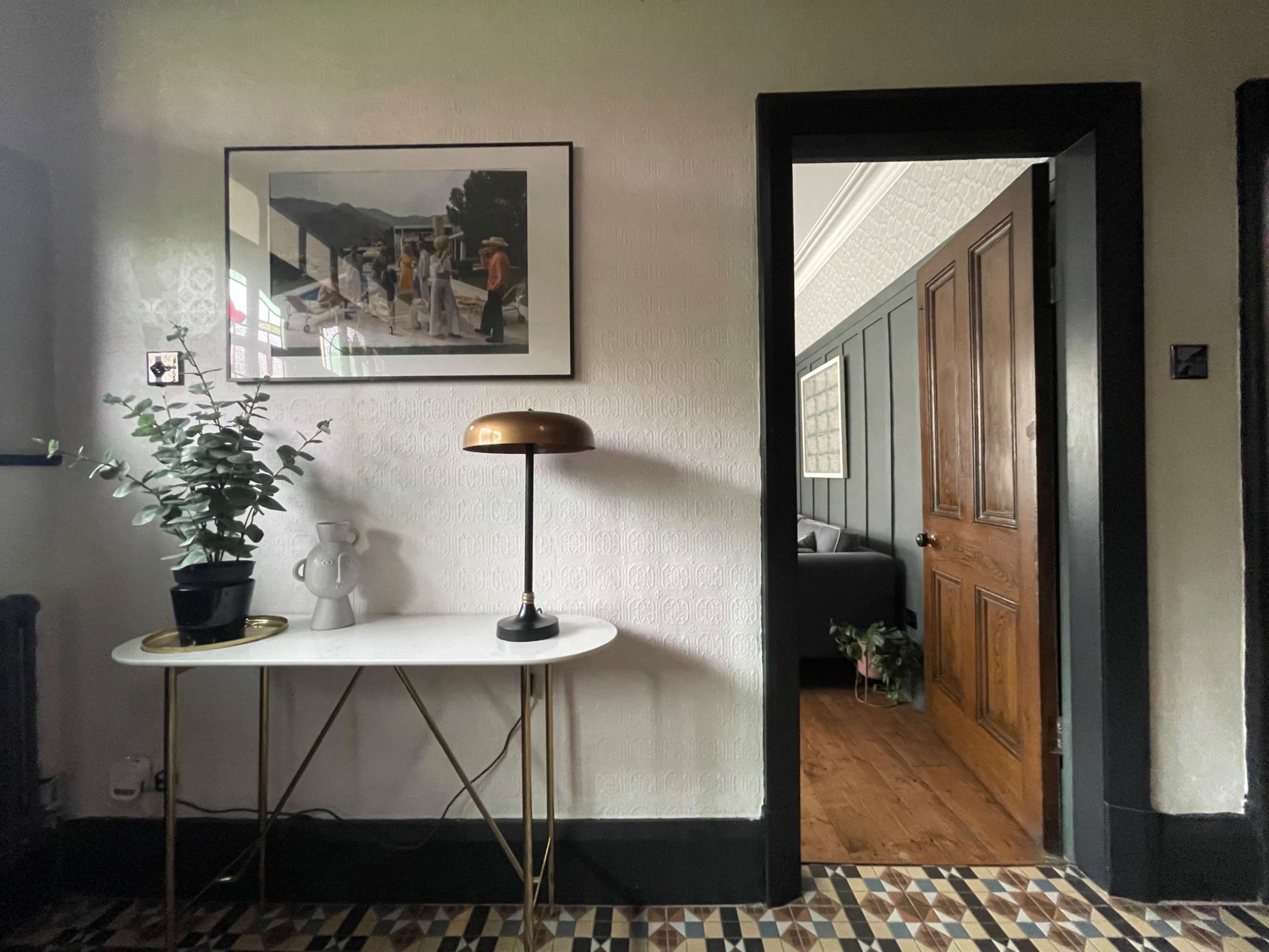 A small entryway features a table with a lamp and a plant, along with a framed picture on the wall and a doorway leading to another room.