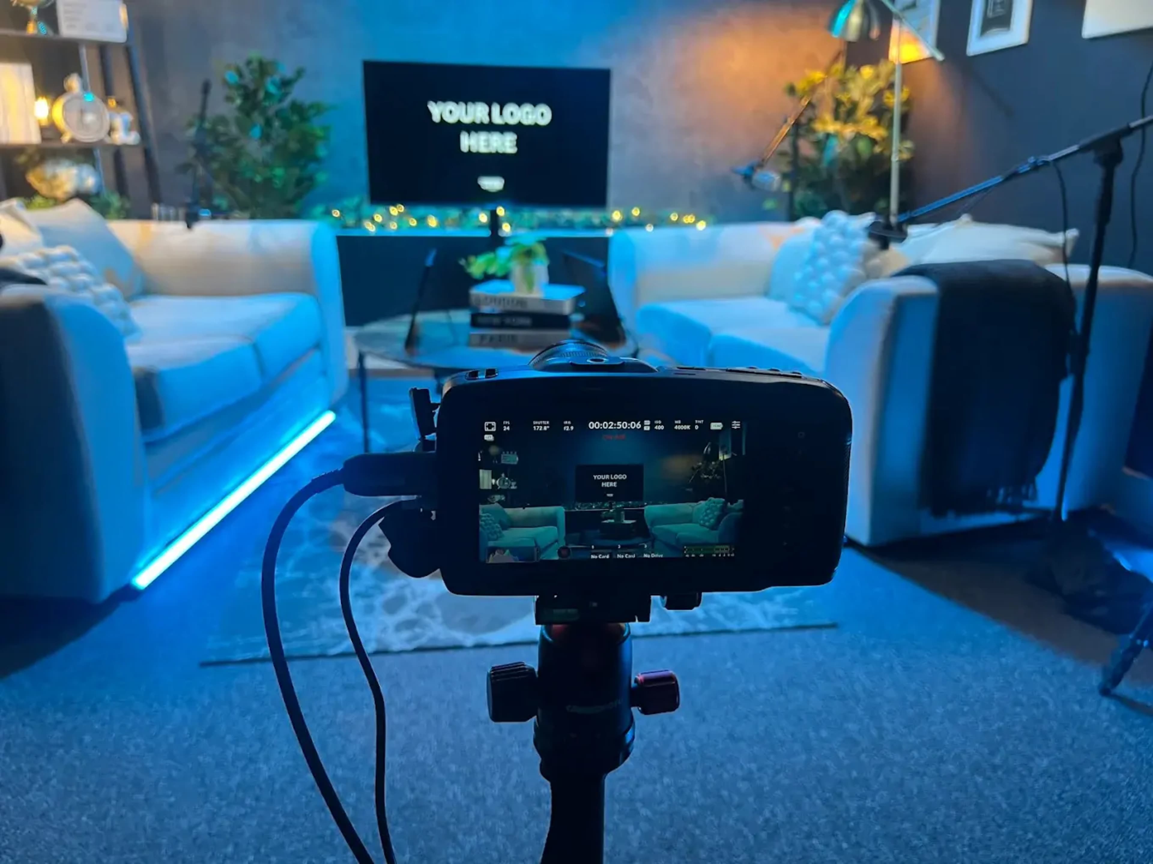 The image shows a camera setup focused on a cozy living room scene with two sofas, a television displaying "YOUR LOGO HERE," and decorative plants under soft lighting.