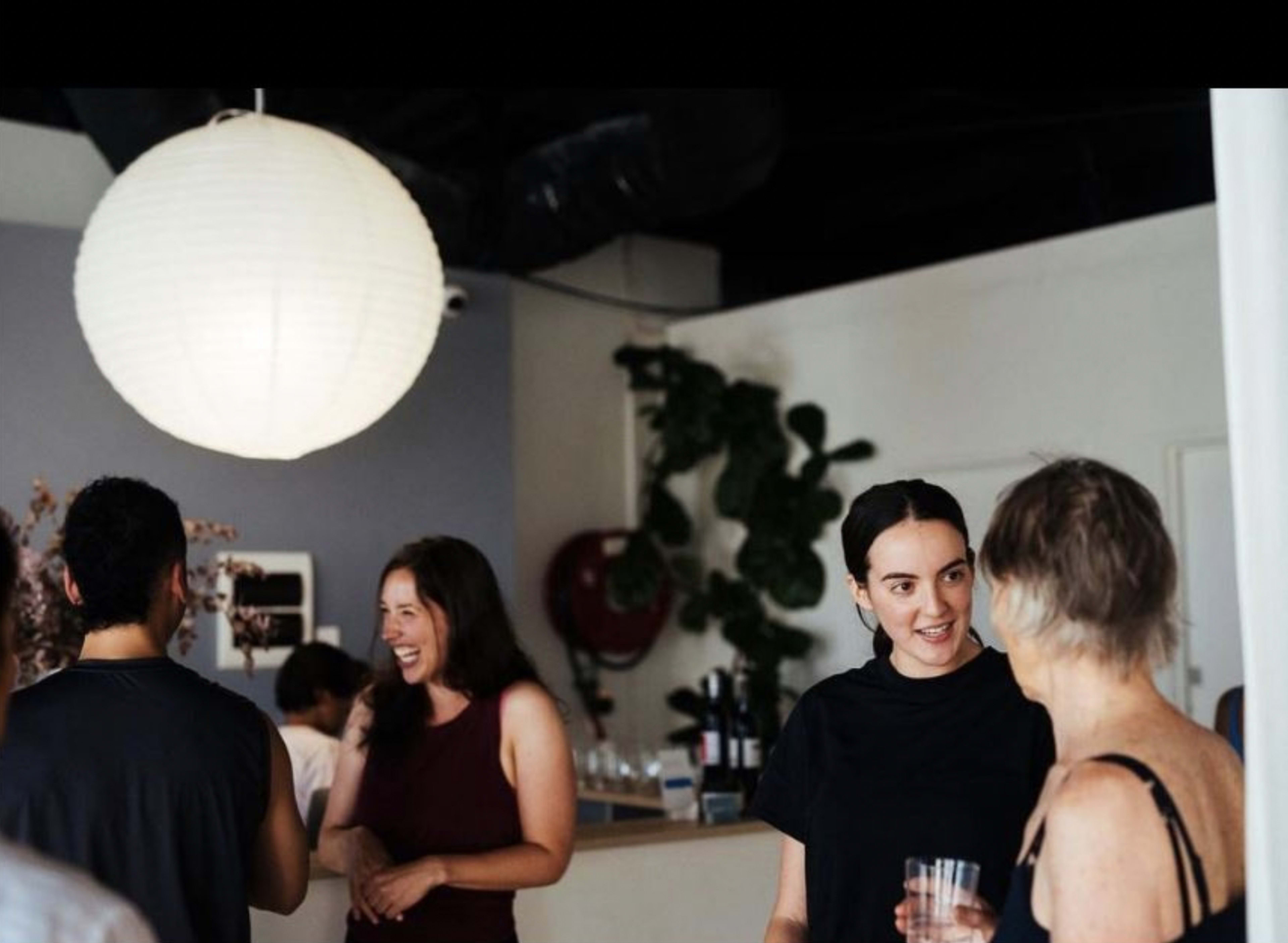 A group of people is engaged in conversation in a modern indoor space with a large paper lantern and potted plants.