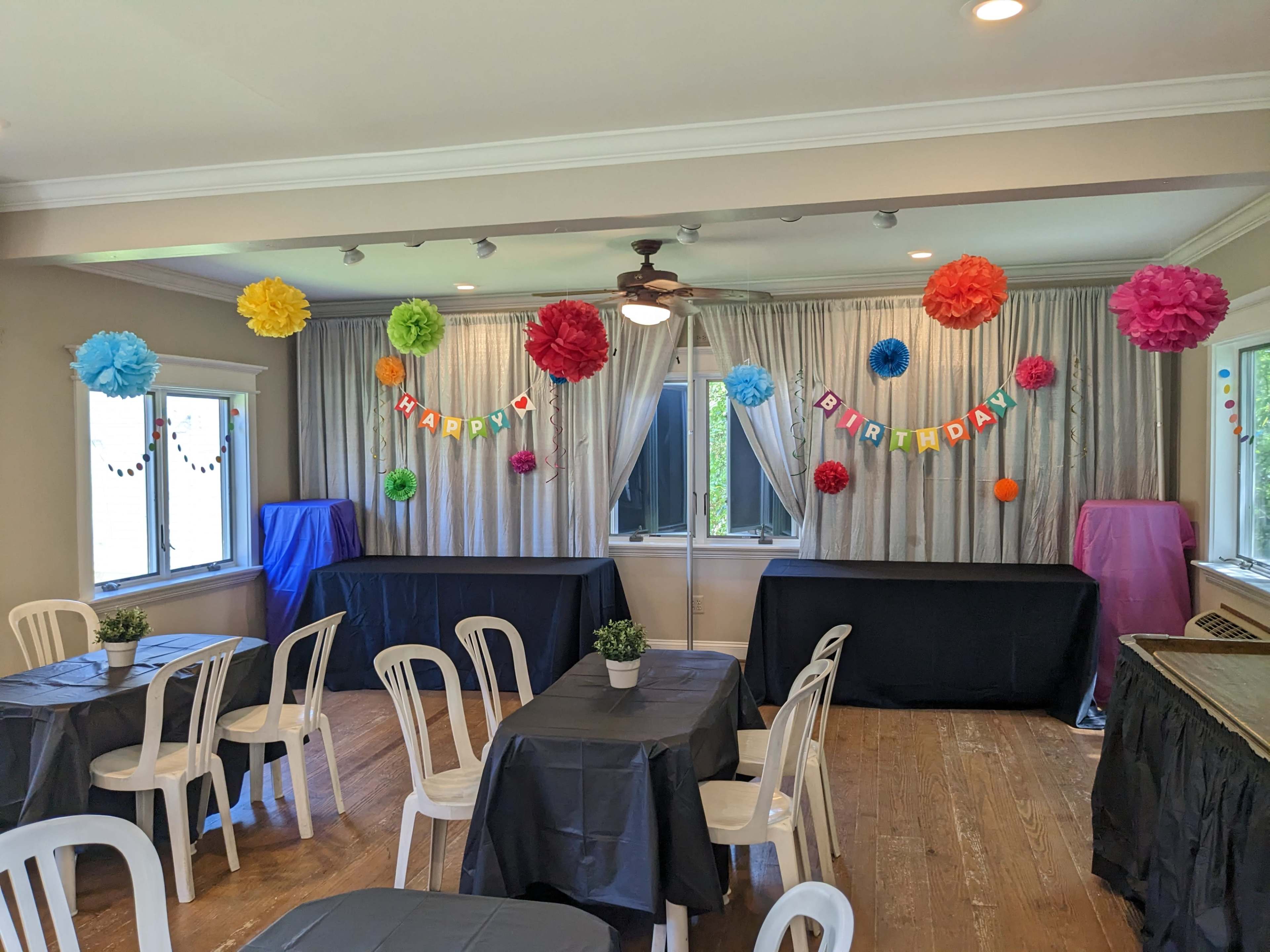 A decorated event space features colorful tissue paper flowers and a "Happy Birthday" banner, with tables and chairs arranged for a celebration.