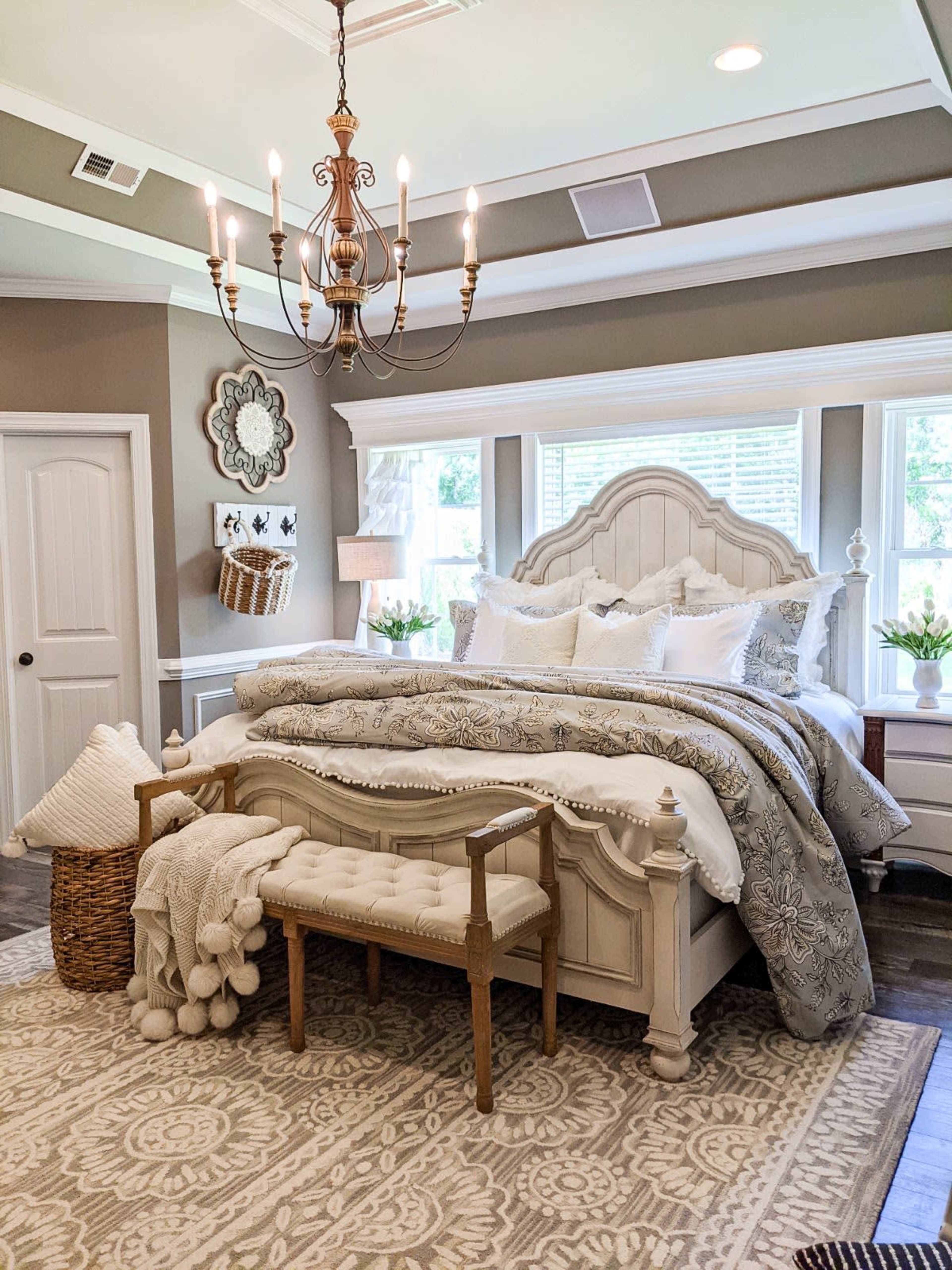The bedroom features a large, upholstered bed with decorative bedding, a bench at the foot, and a chandelier overhead, all set against a neutral-colored wall.