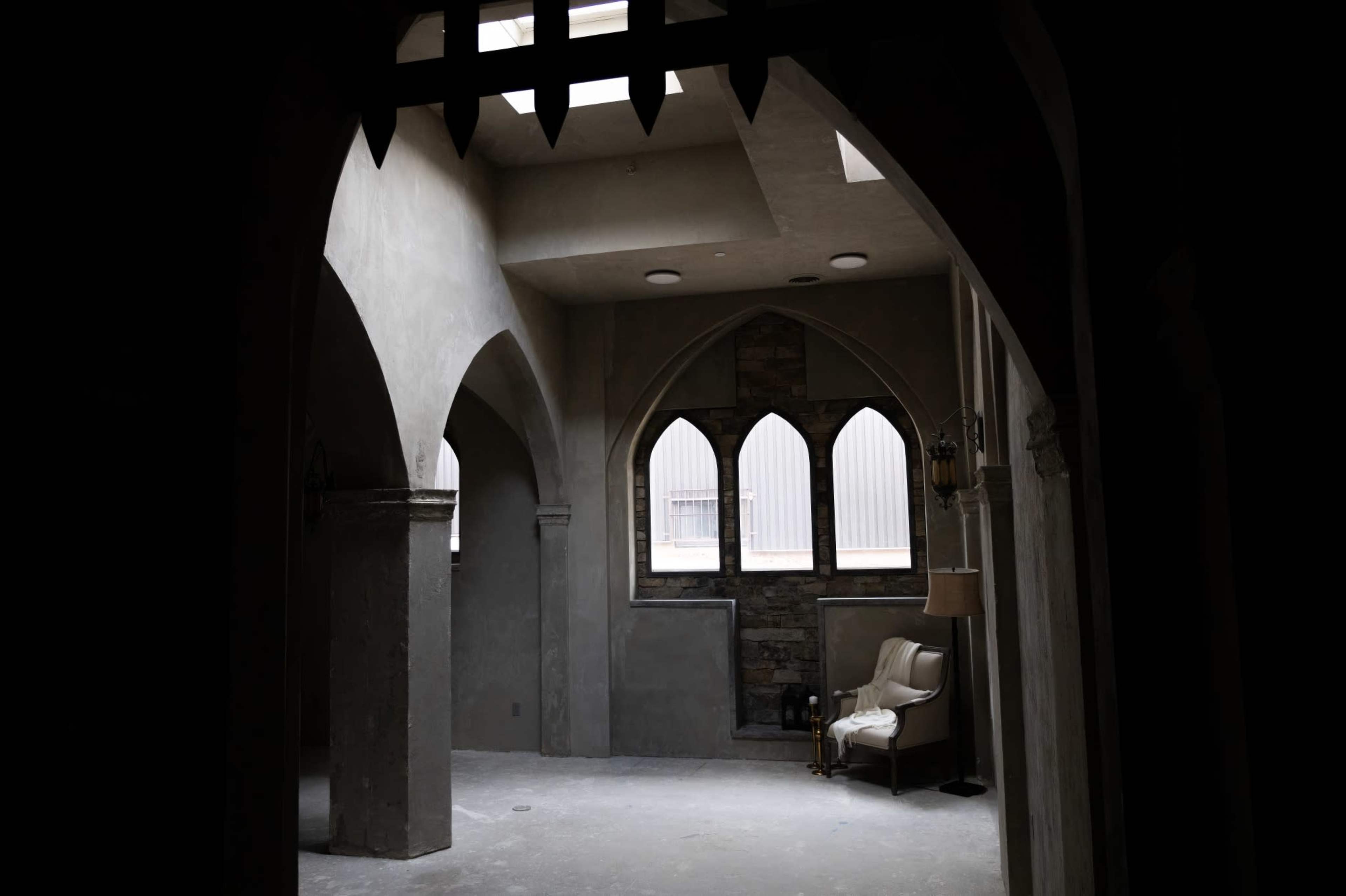 The image shows an empty room with arched doorways and windows, featuring a single chair and a stone wall accent.