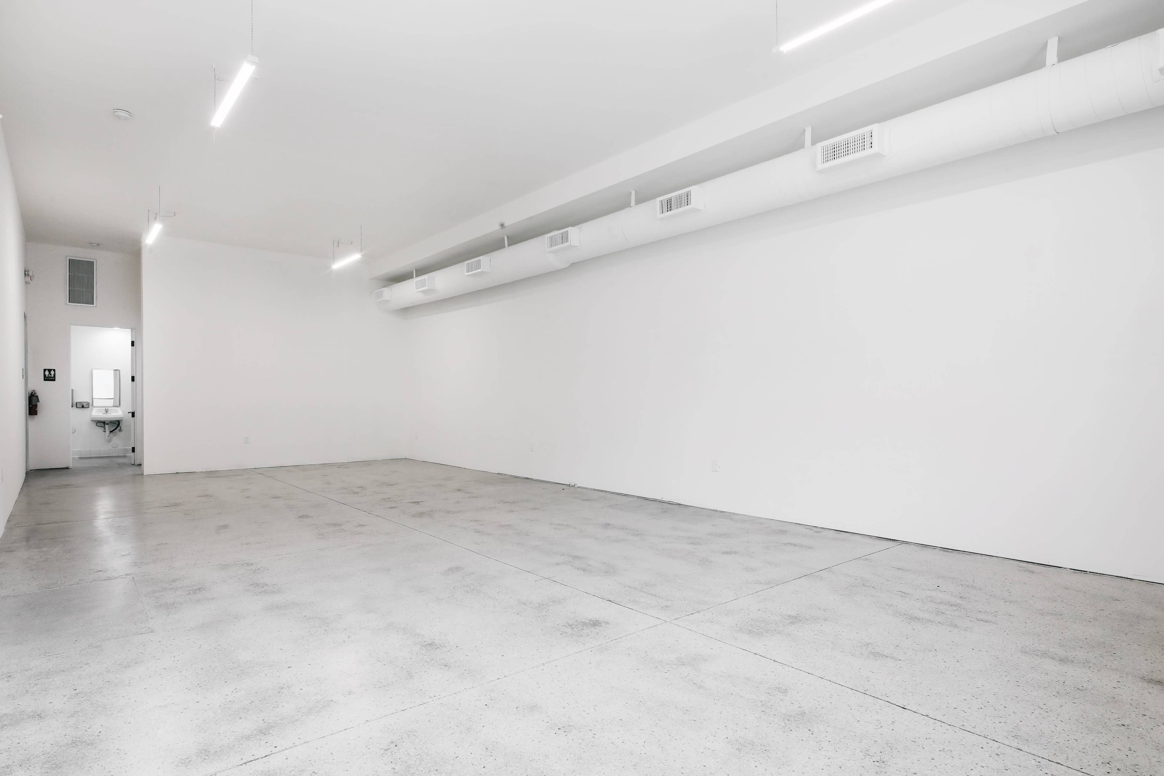 A spacious, empty room with concrete flooring and white walls, featuring fluorescent lighting and air ducts along the ceiling.