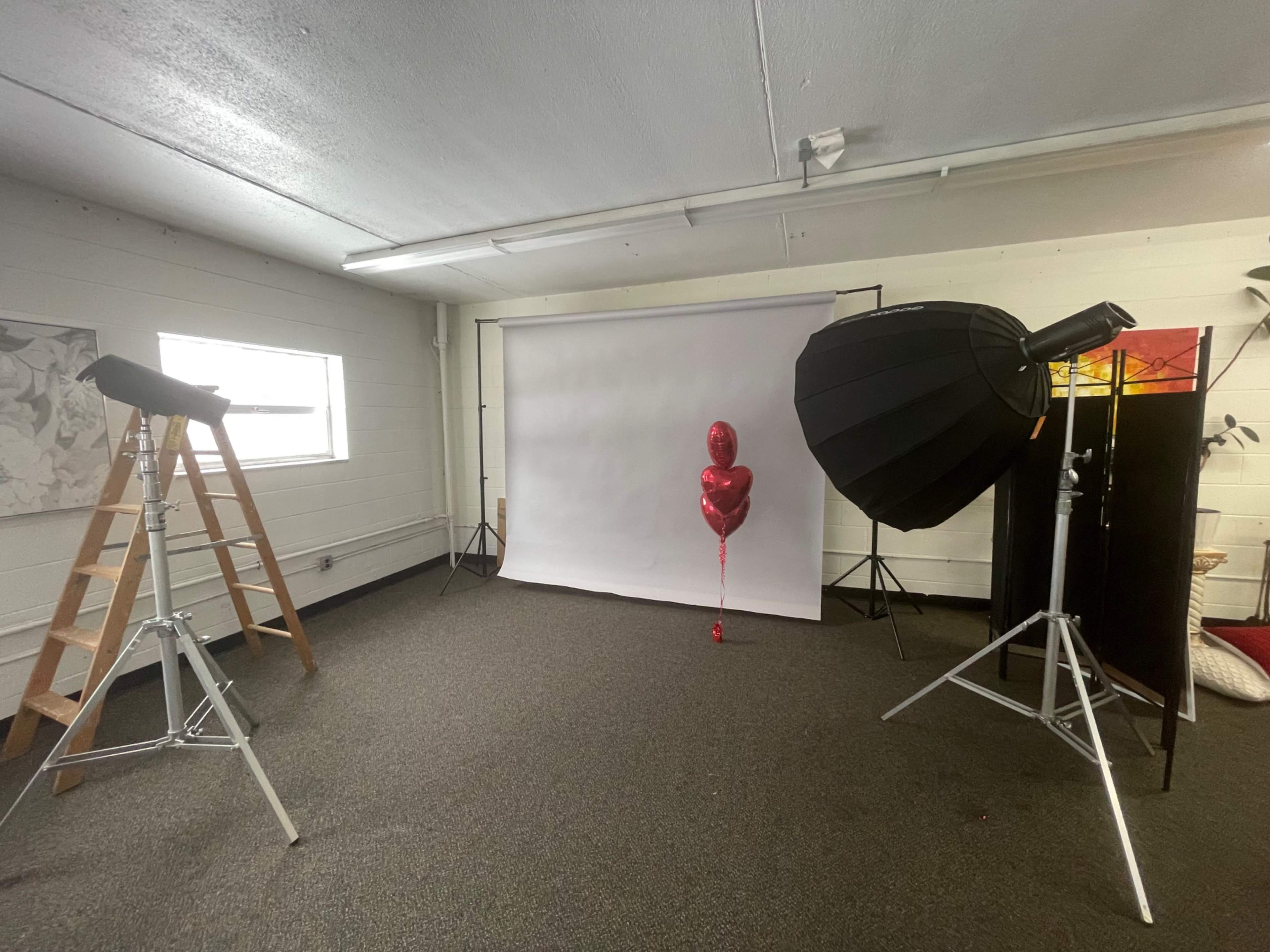 A photography studio is set up with a backdrop, lighting equipment, a ladder, and a single red balloon on a stand.