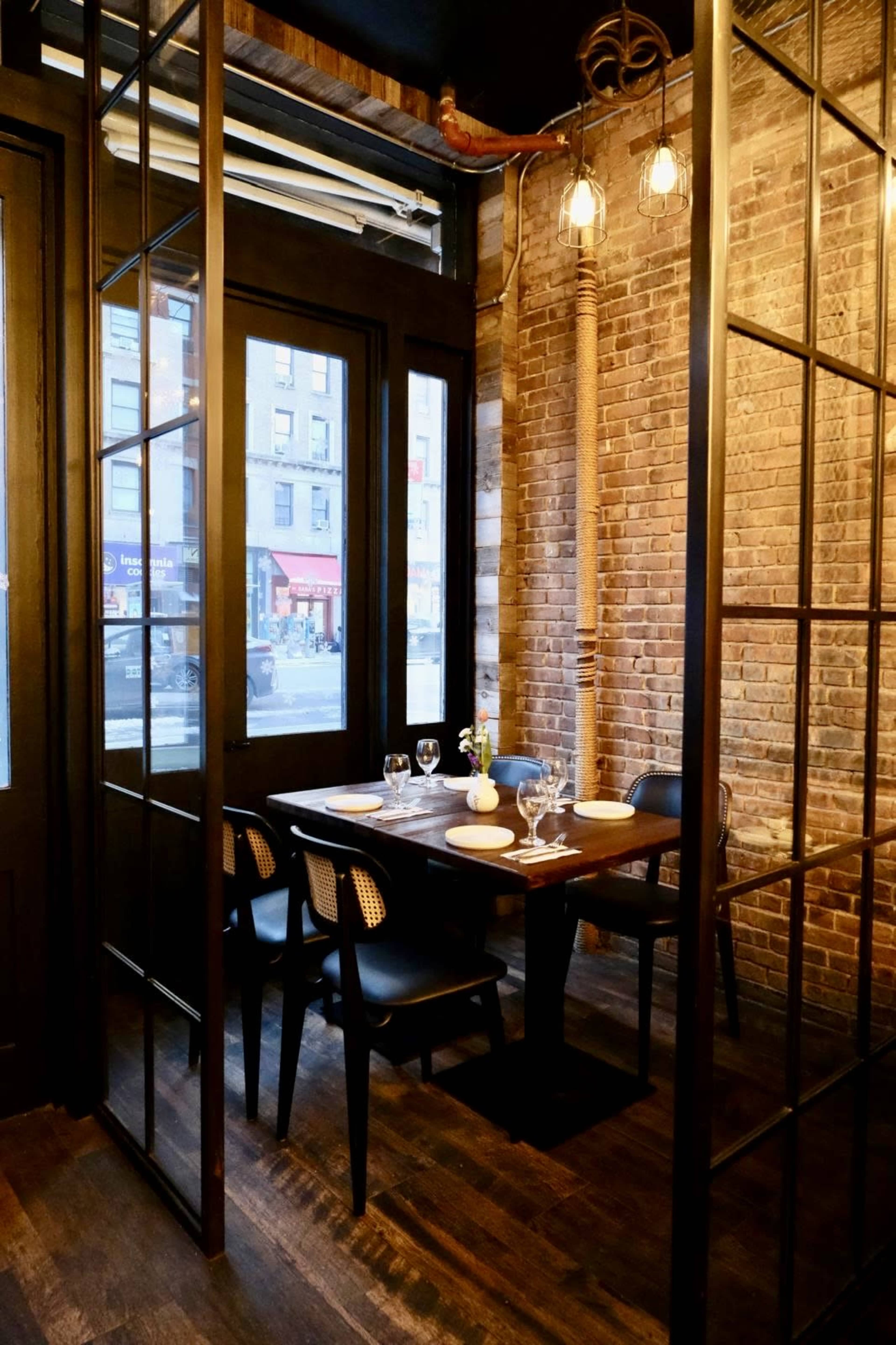 A cozy dining area features a wooden table set for four, enclosed by glass and metal partitions against a backdrop of exposed brick walls.
