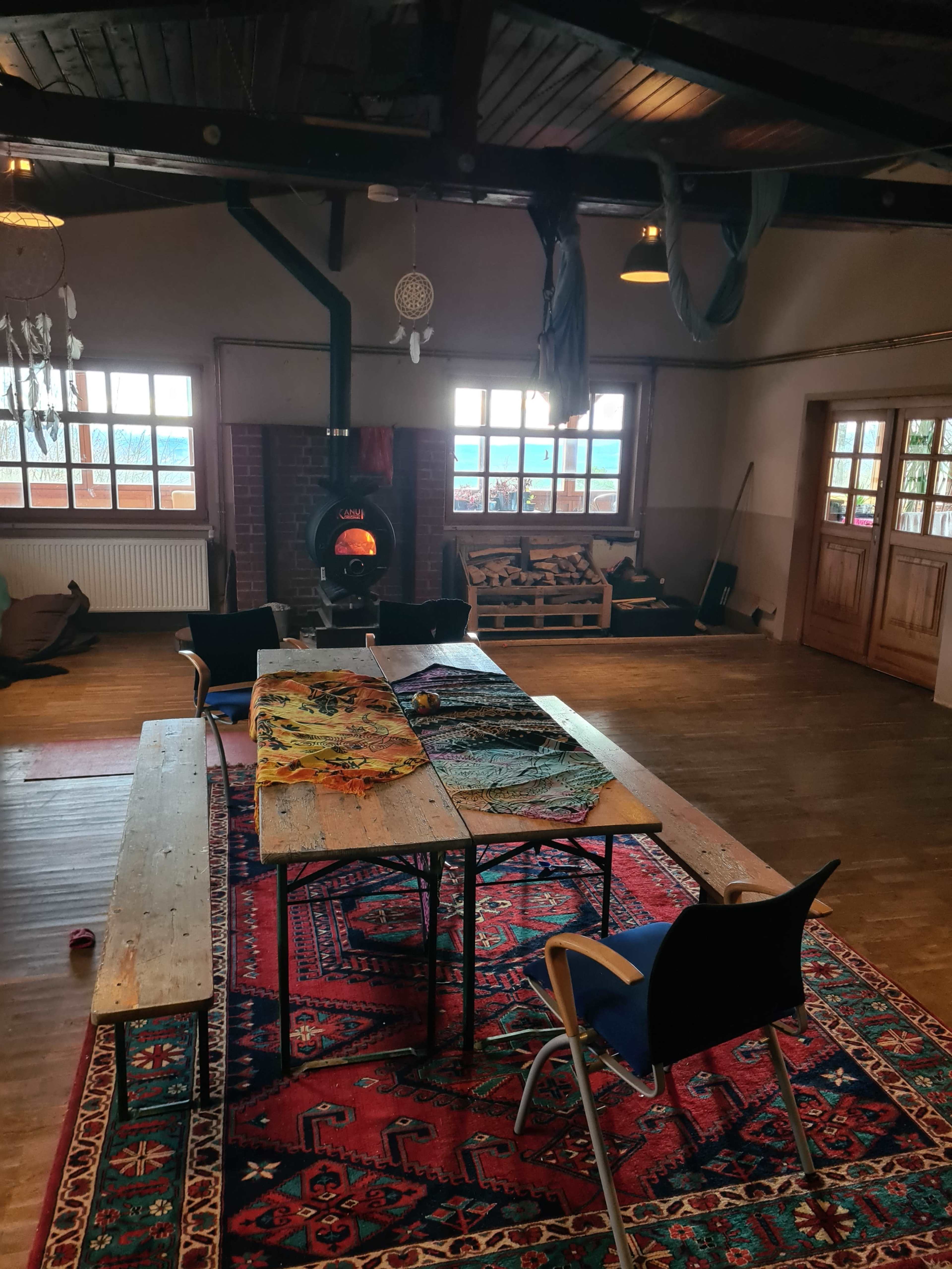 A spacious room features a wooden floor, a large patterned rug, a fireplace, and several tables and chairs arranged for a gathering.