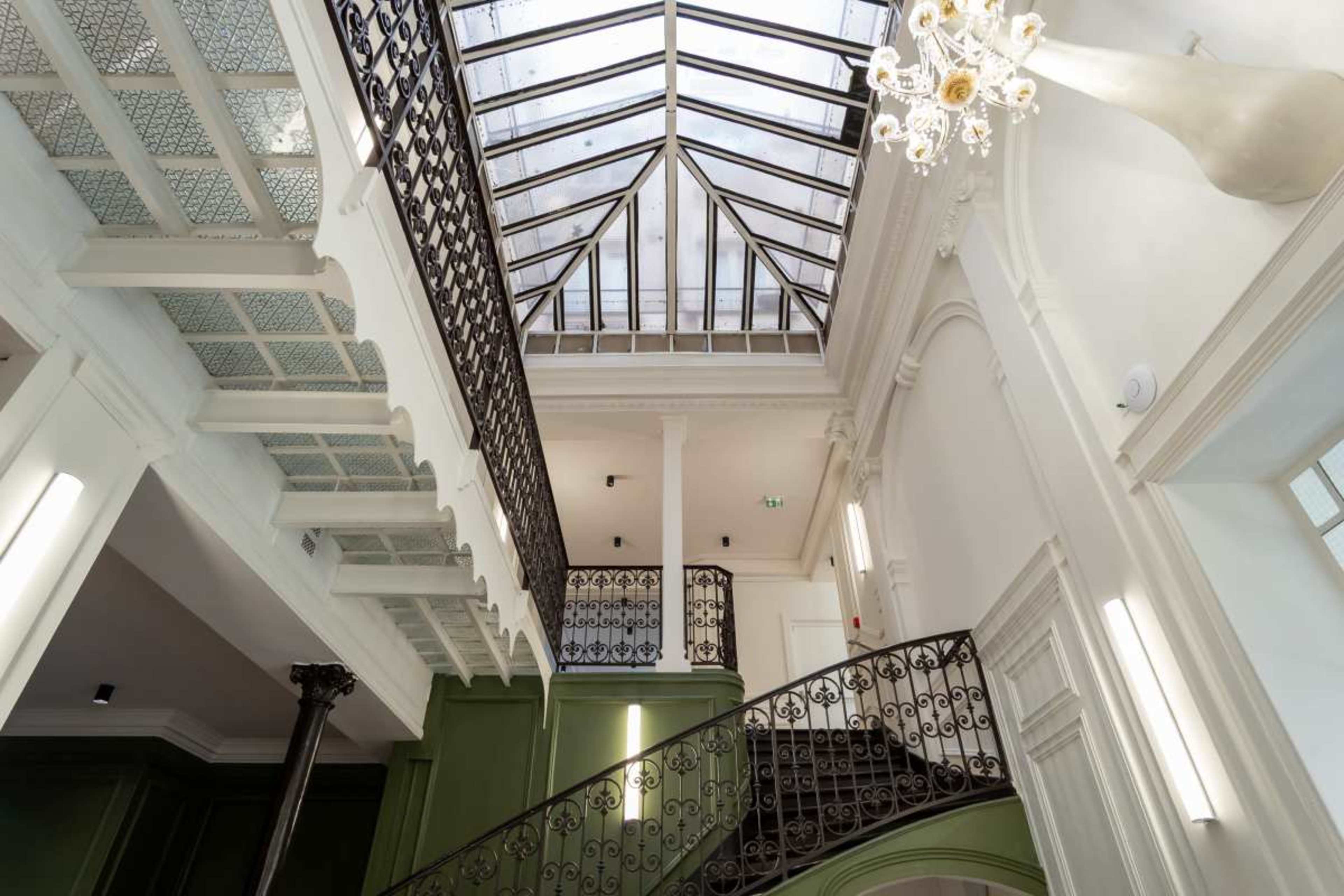 The image shows a spacious interior with a grand staircase, wrought iron railing, and a ceiling featuring large glass panels.
