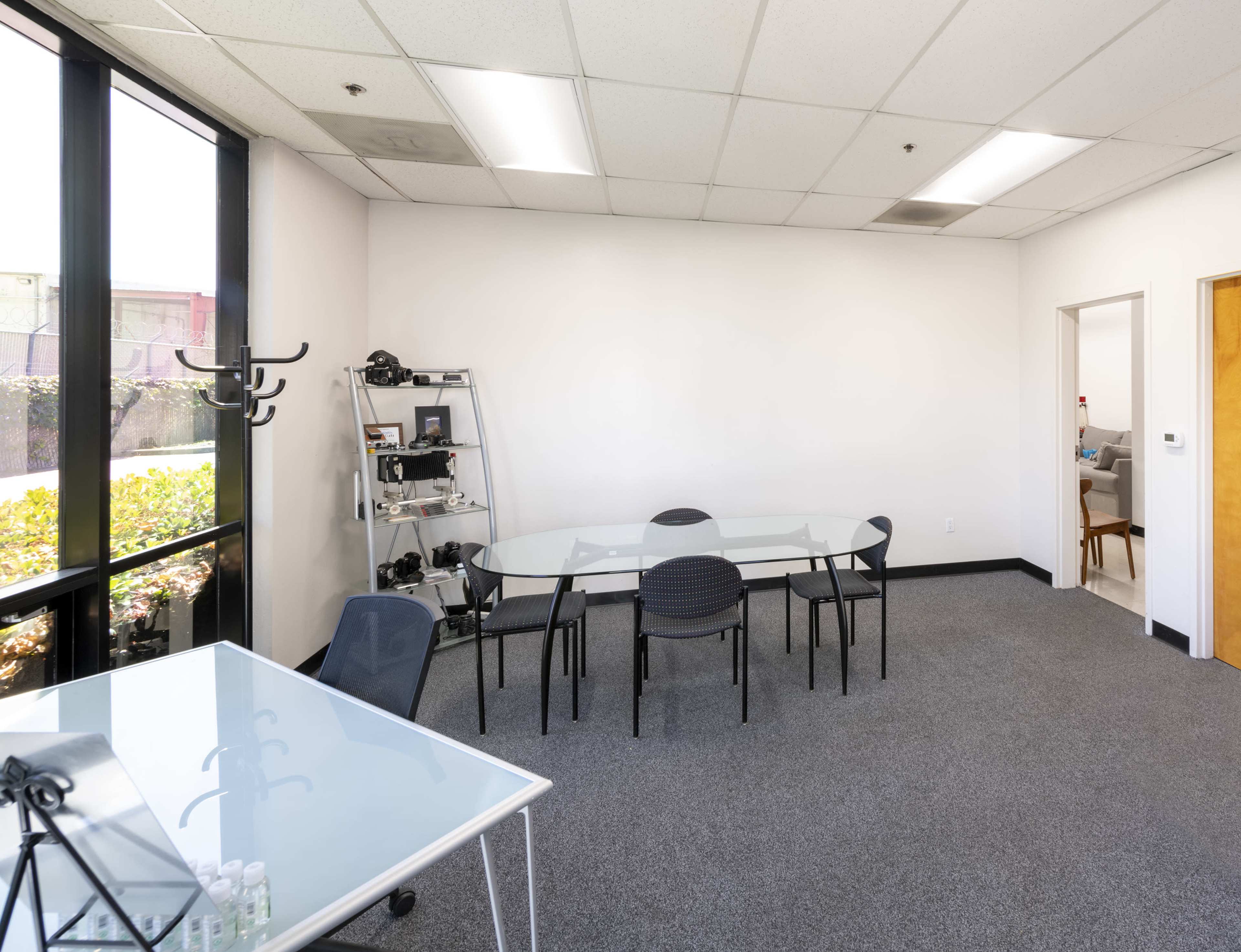 A bright office space with glass tables, black chairs, and a shelf displaying various items, alongside a doorway leading to another room.